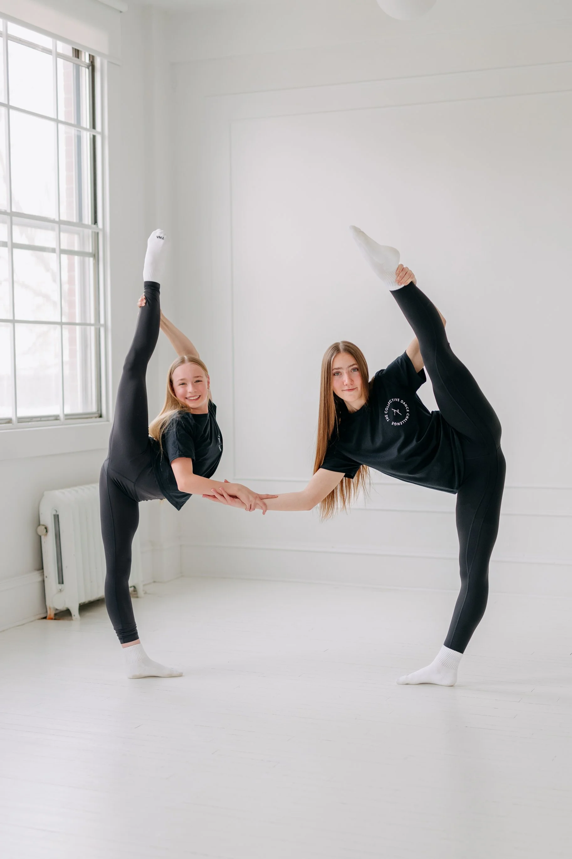 Two young girls perform a balancing yoga pose together in a bright, minimalist dance studio with white walls and large windows.