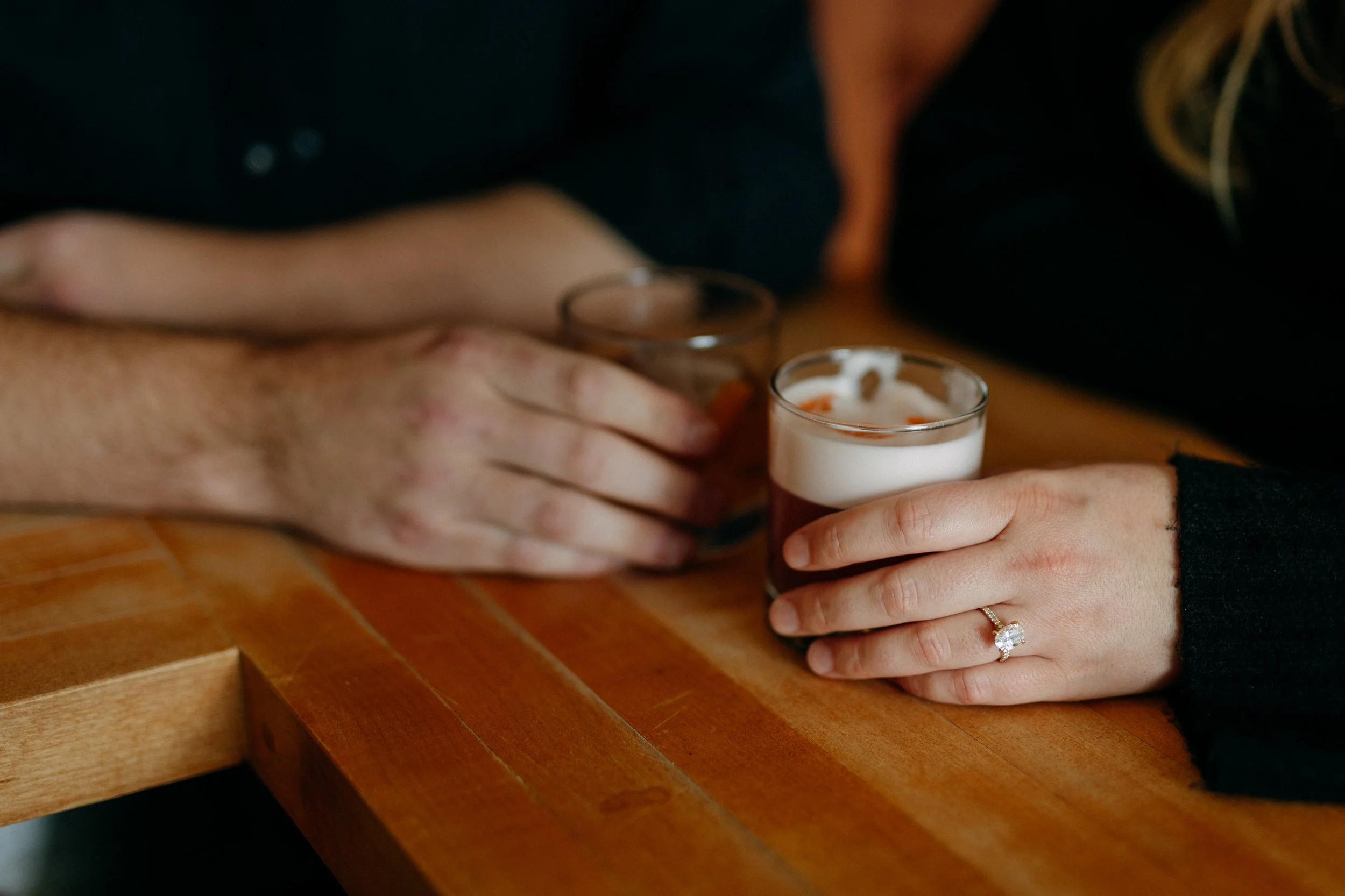 Two people holding glasses of drinks on a wooden table, one with an engagement ring visible.