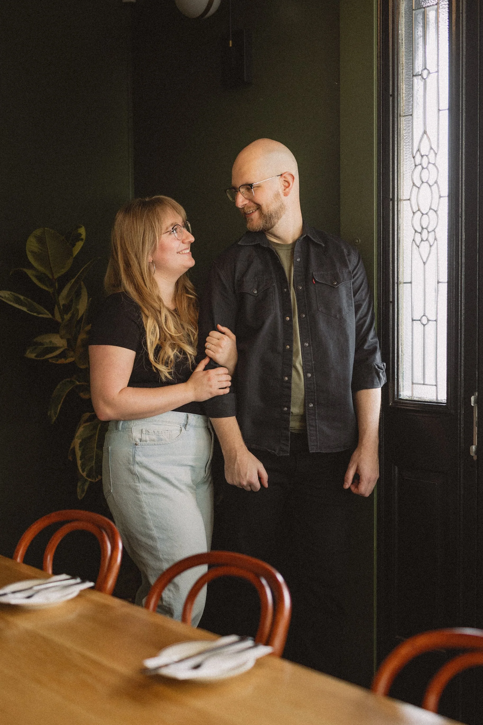 A man and a woman standing close together, smiling and looking at each other indoors near a window with decorative glass.