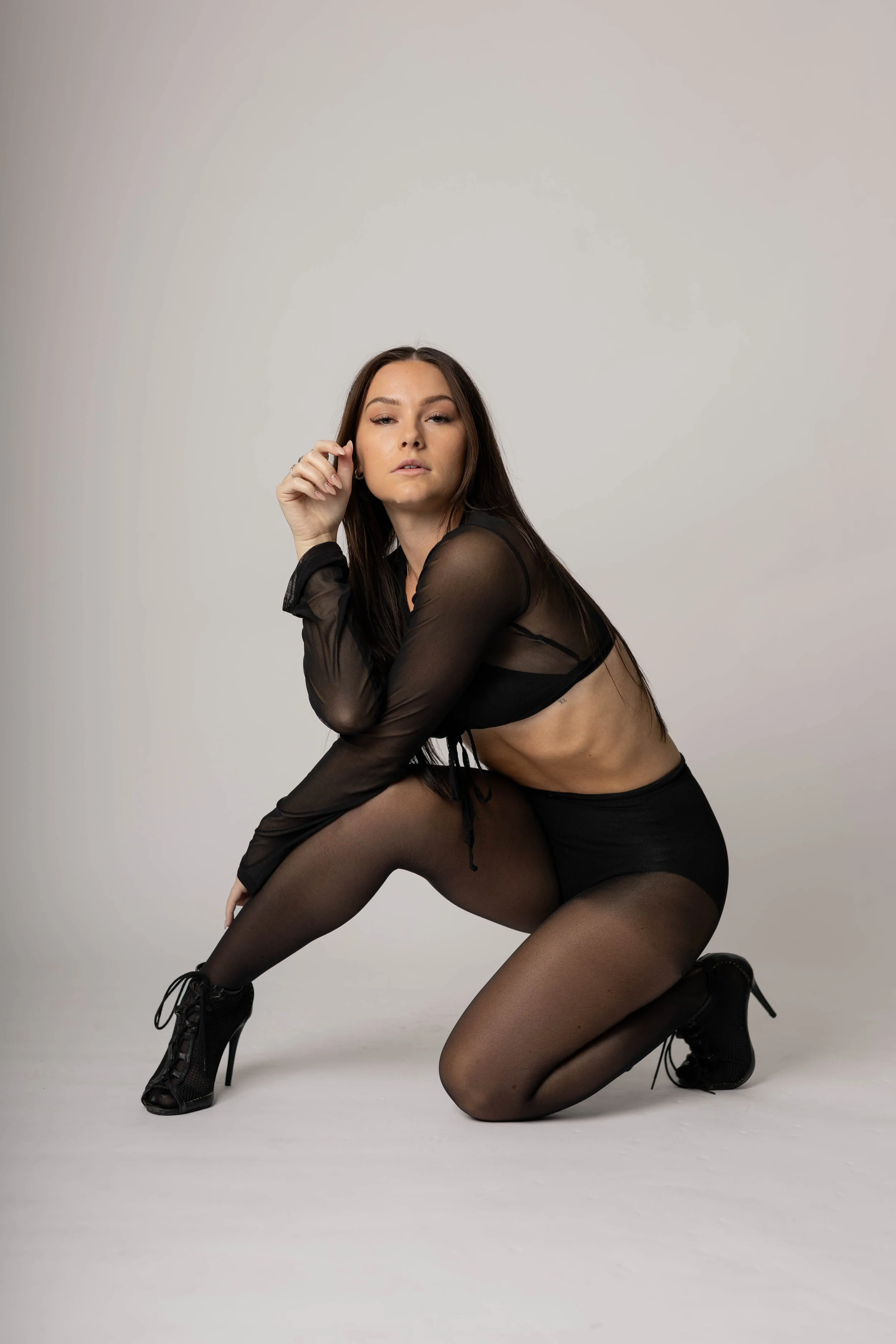 A woman in black sheer clothing, kneeling on the floor with one leg bent and the other extended, against a plain white background.