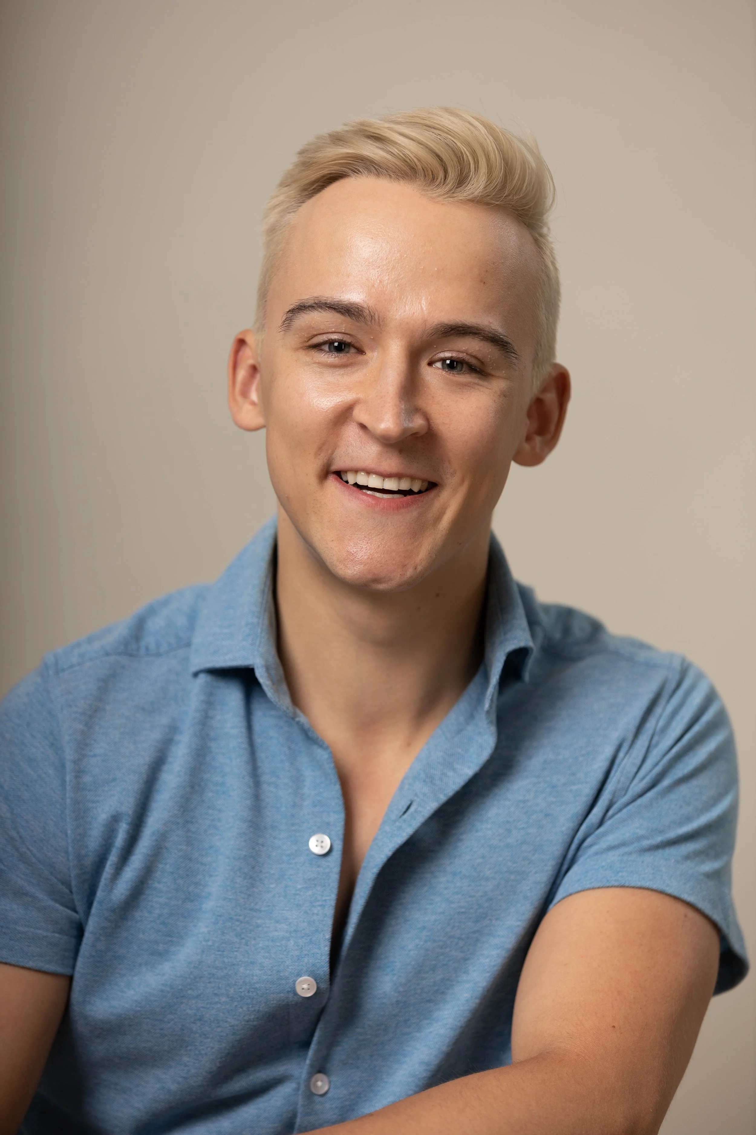 A young man with short blond hair, wearing a light blue button-up shirt, smiling and looking to his left against a plain background.