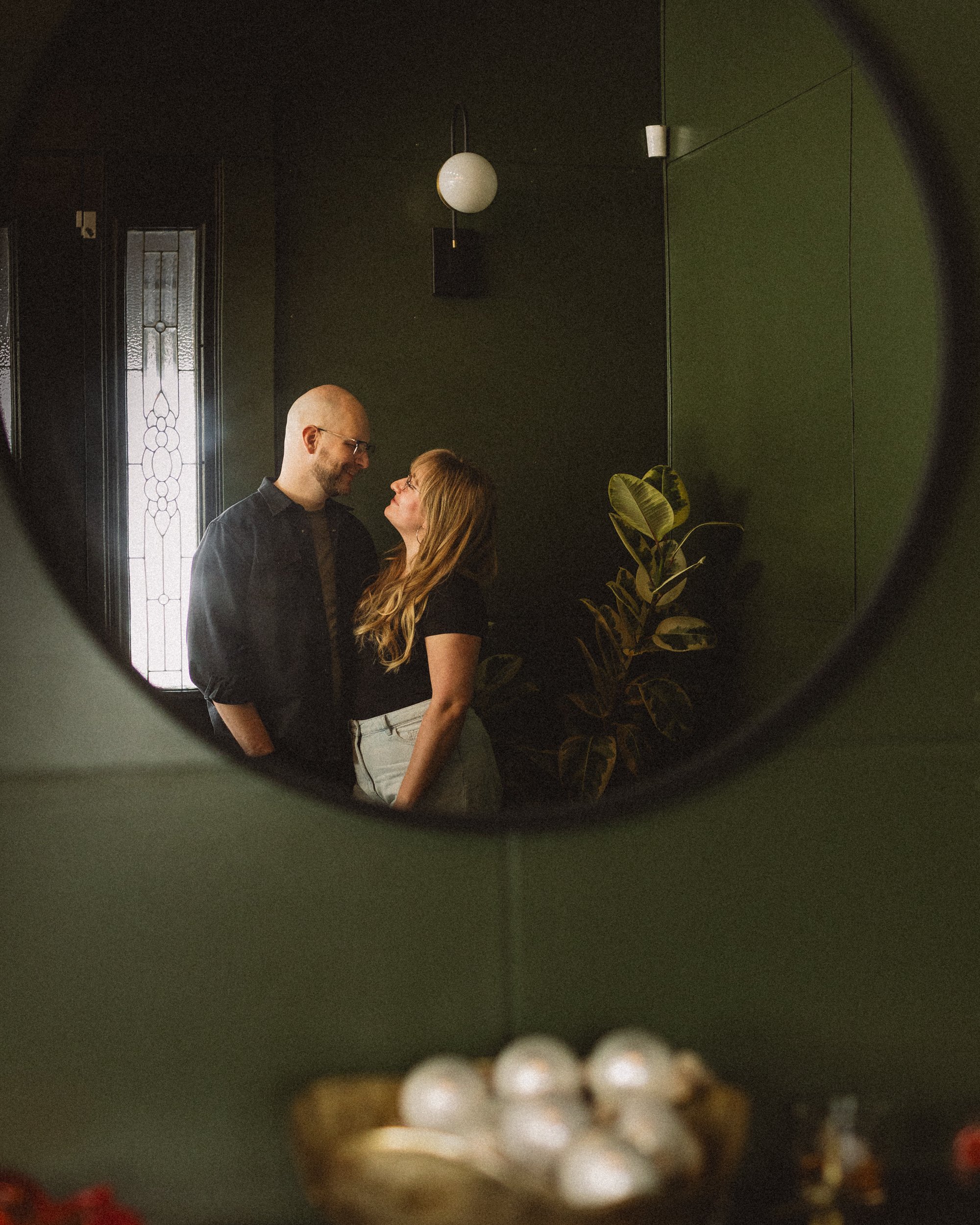 A couple looking at each other and smiling, seen through a circular mirror with a green wall, a plant, a window, and a light fixture in the background.