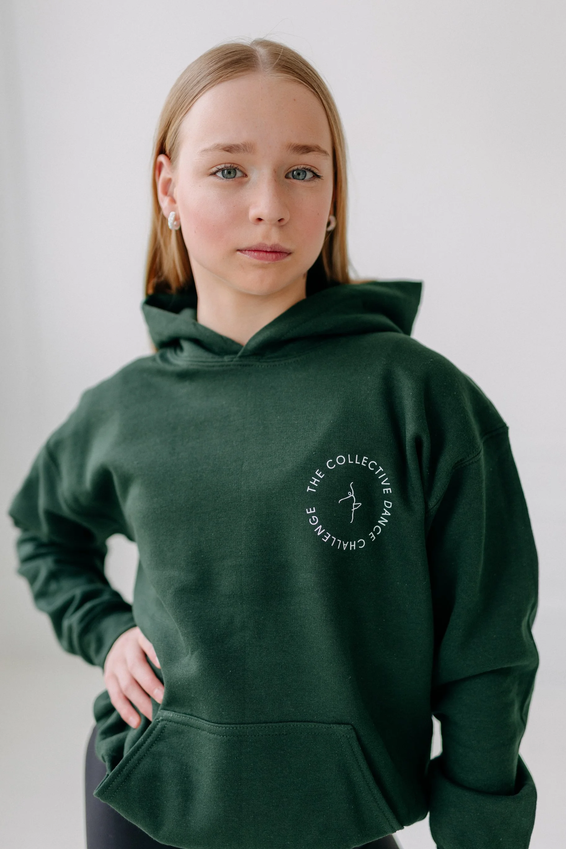 A young woman with blonde hair and light skin, wearing a dark green hoodie with a white circular logo that says "The Collective Dance Change" around a stylized dancer icon. She is standing against a plain light background, with her right hand on her 