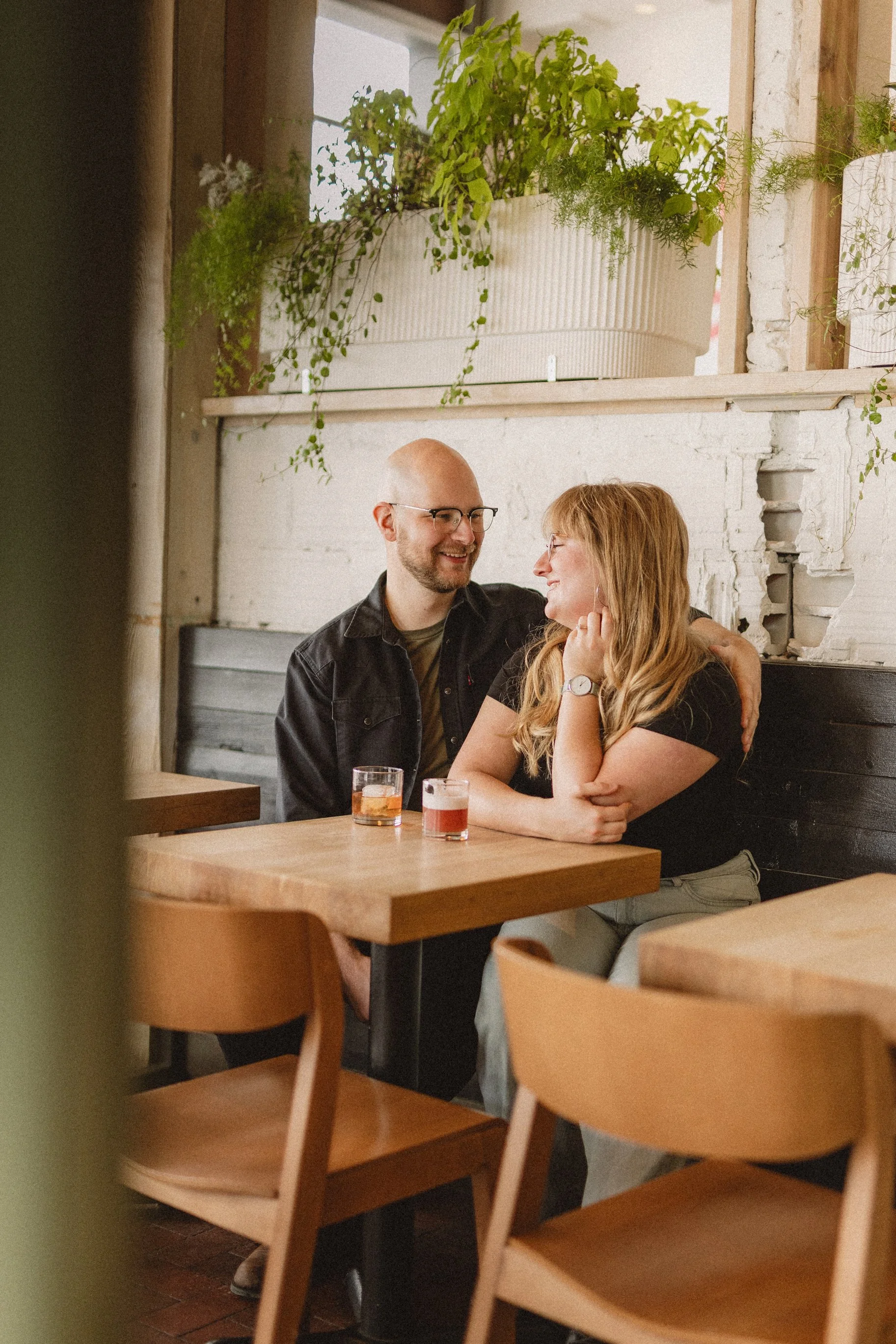 A couple enjoying drinks at a wooden table in a cozy cafe with white brick walls and potted plants above
