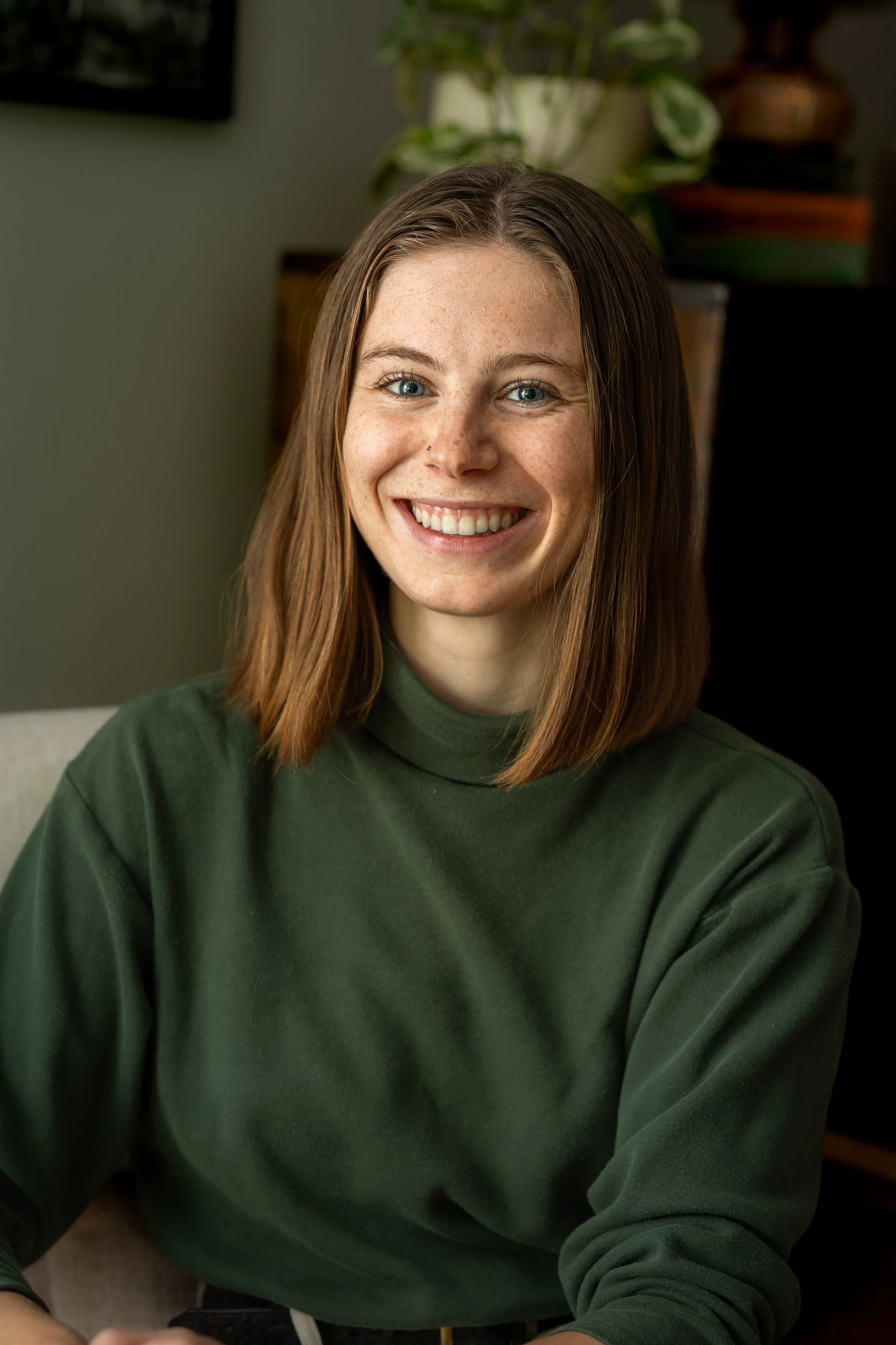 A smiling young woman with shoulder-length brown hair, wearing a green sweatshirt, sitting indoors with a blurred background of plants and furniture.