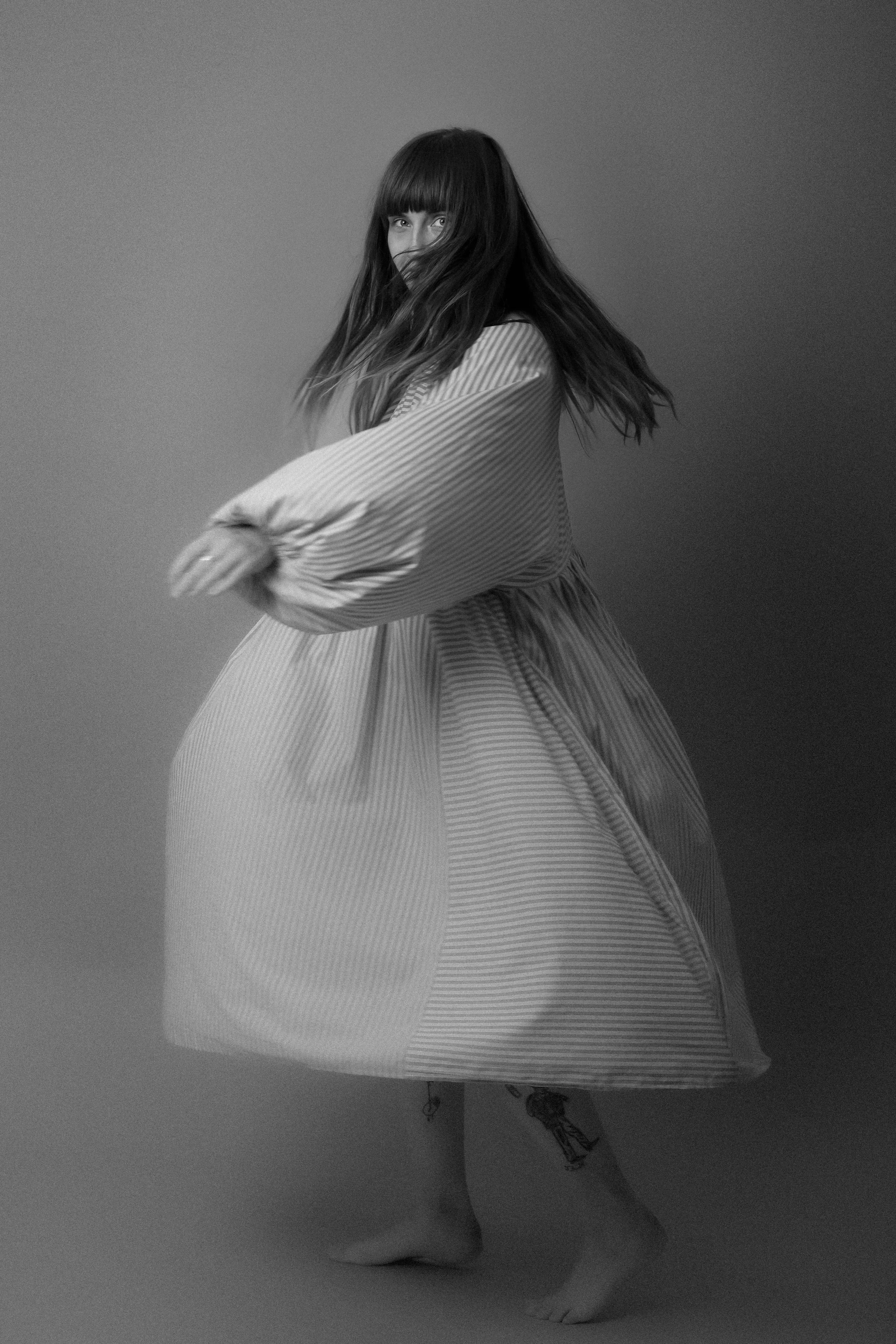 A woman with long hair wearing a striped dress, similar in appearance to a kimono, standing on one foot and turning to the side against a plain background.