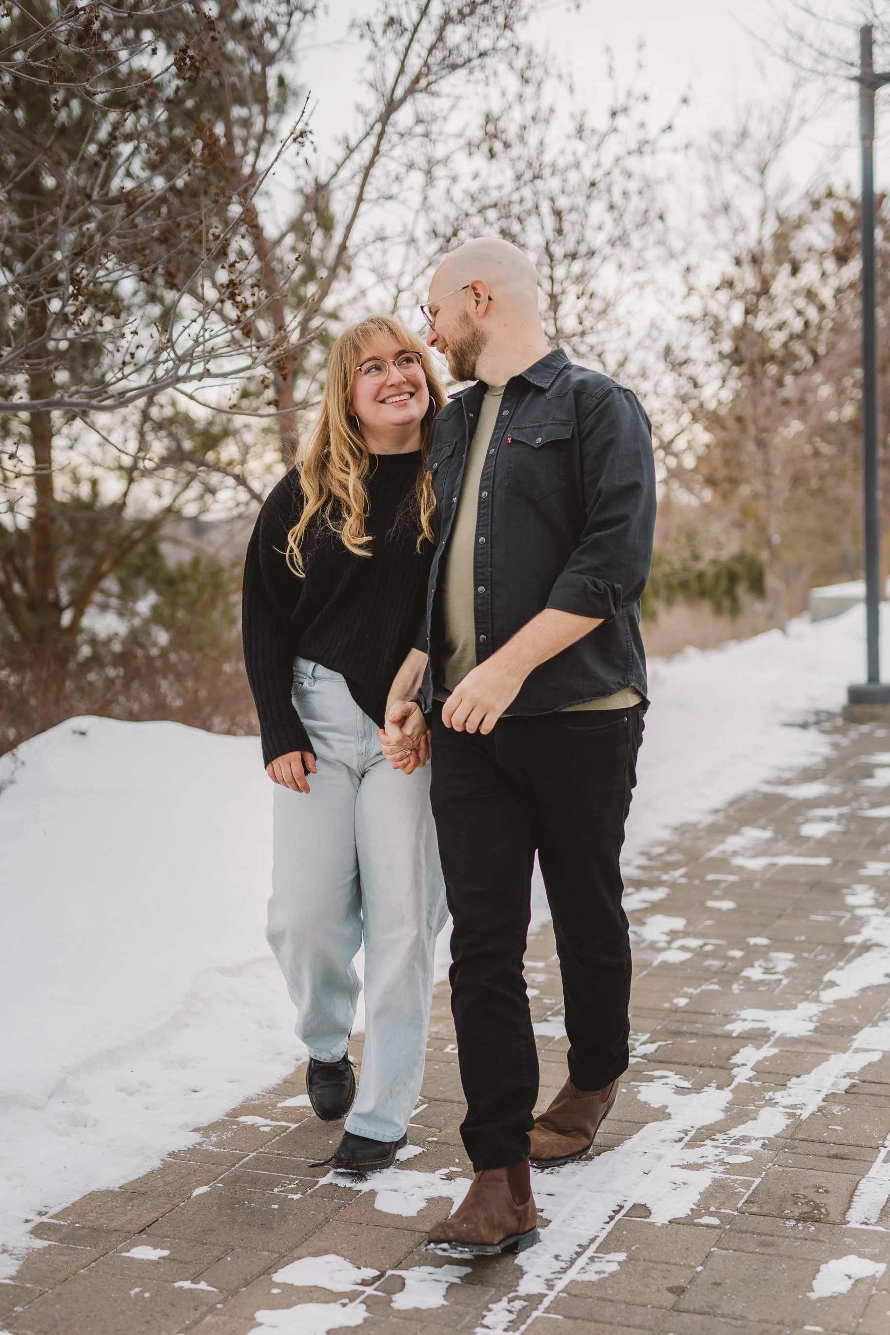 A couple walking hand in hand outdoors during winter, surrounded by snow and leafless trees, smiling at each other.