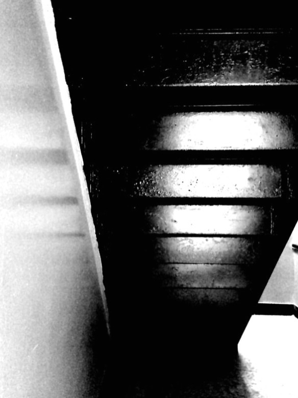 Close-up of a dark staircase with horizontal treads, viewed from the bottom looking upward, in black and white.