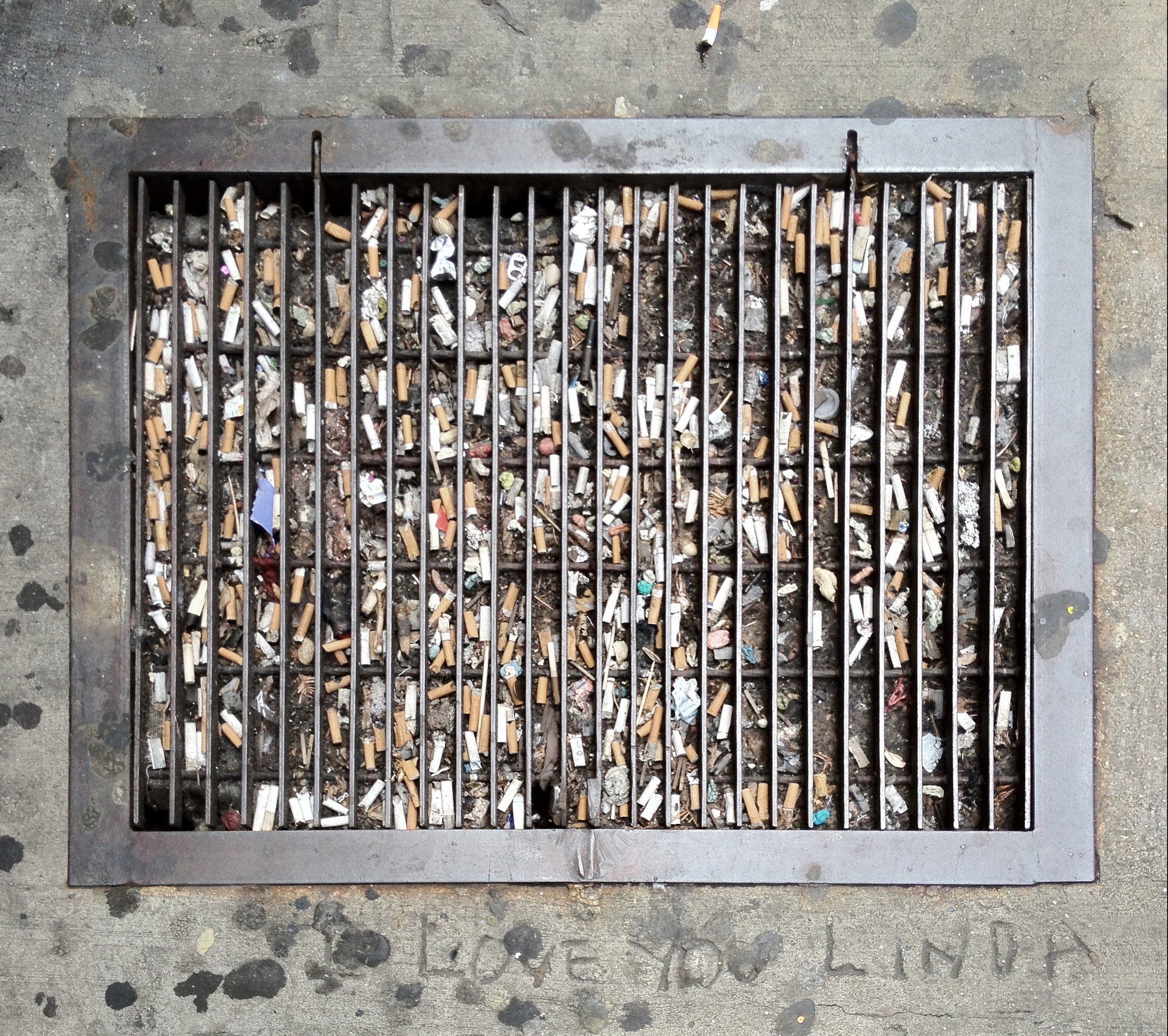 A metal street grate filled with cigarette butts, small pieces of trash, and debris on a concrete sidewalk.