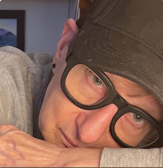 Close-up of a person with glasses and a black cap, resting their head on their crossed arms, looking into the camera.