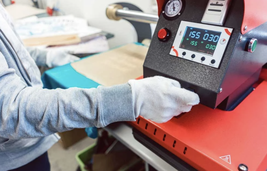 Person wearing gloves operating a heat press machine with digital controls in a workshop.