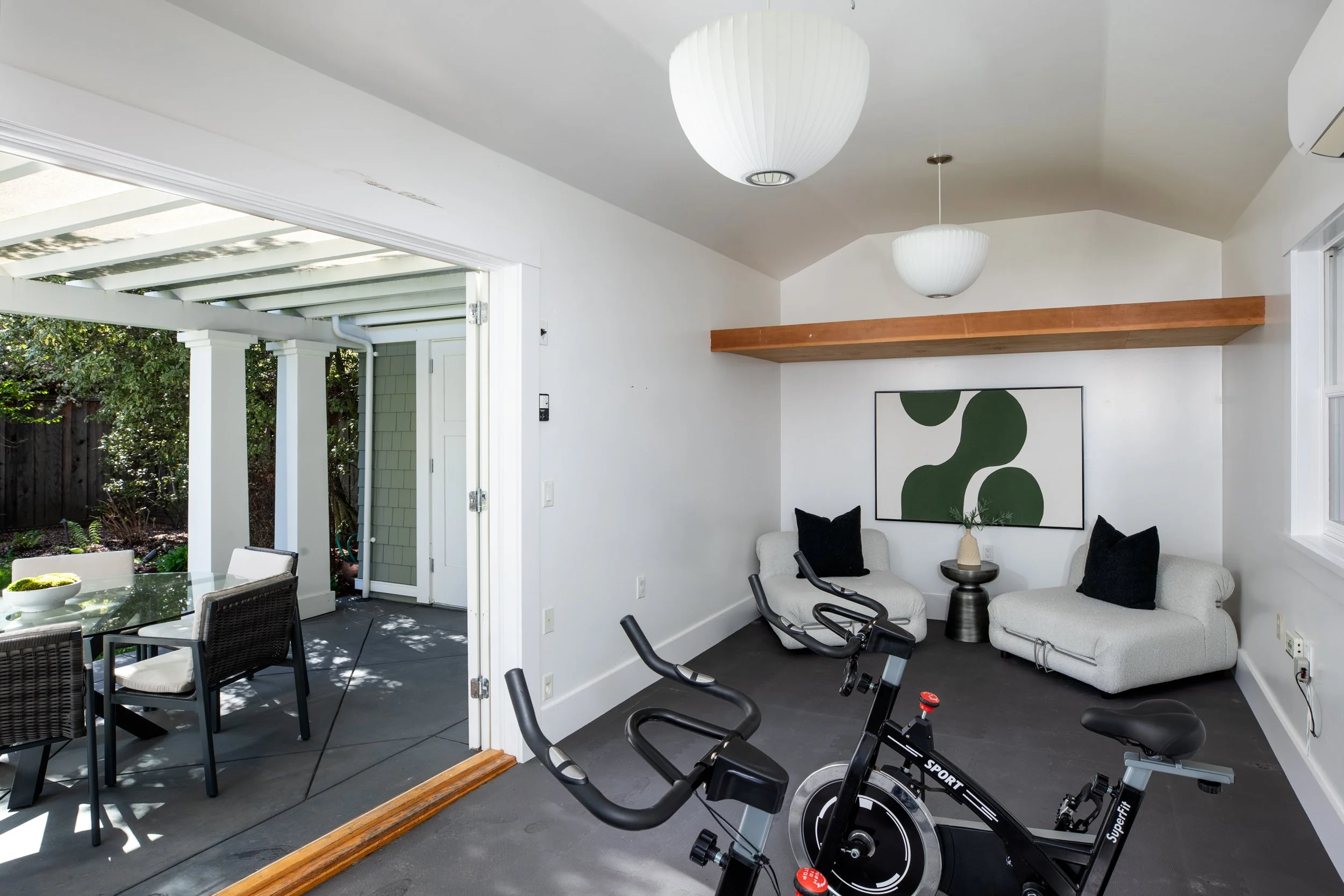 Indoor space with two white couches, black pillows, a small round table with a beige vase and plant, exercise bike, and outdoor patio with dining table and chairs visible through open sliding door.