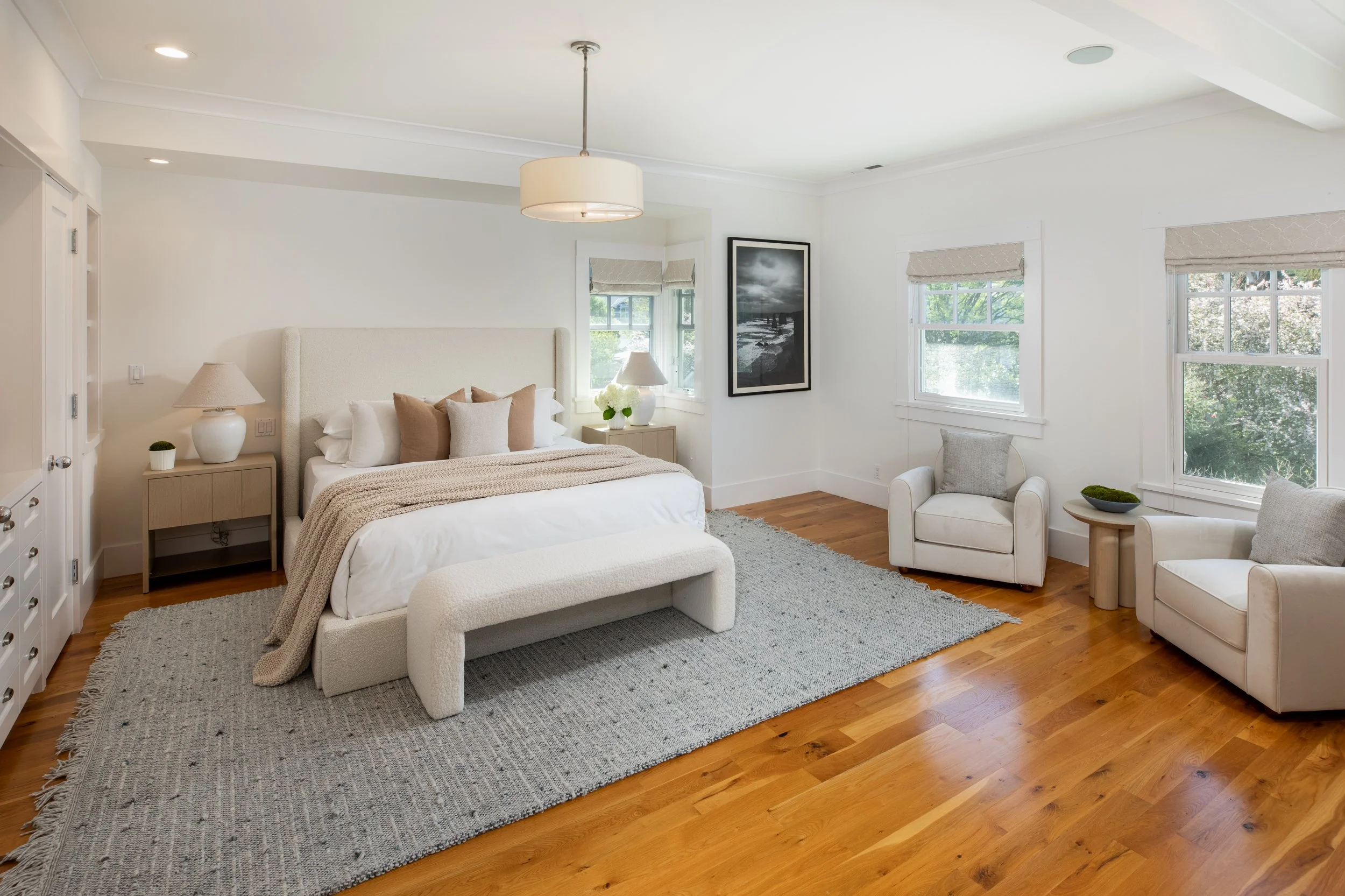 Bright bedroom with white walls, large bed with beige and white bedding, two beige nightstands with lamps, two white armchairs with pillows, wooden flooring, gray rug, and large windows with blinds.