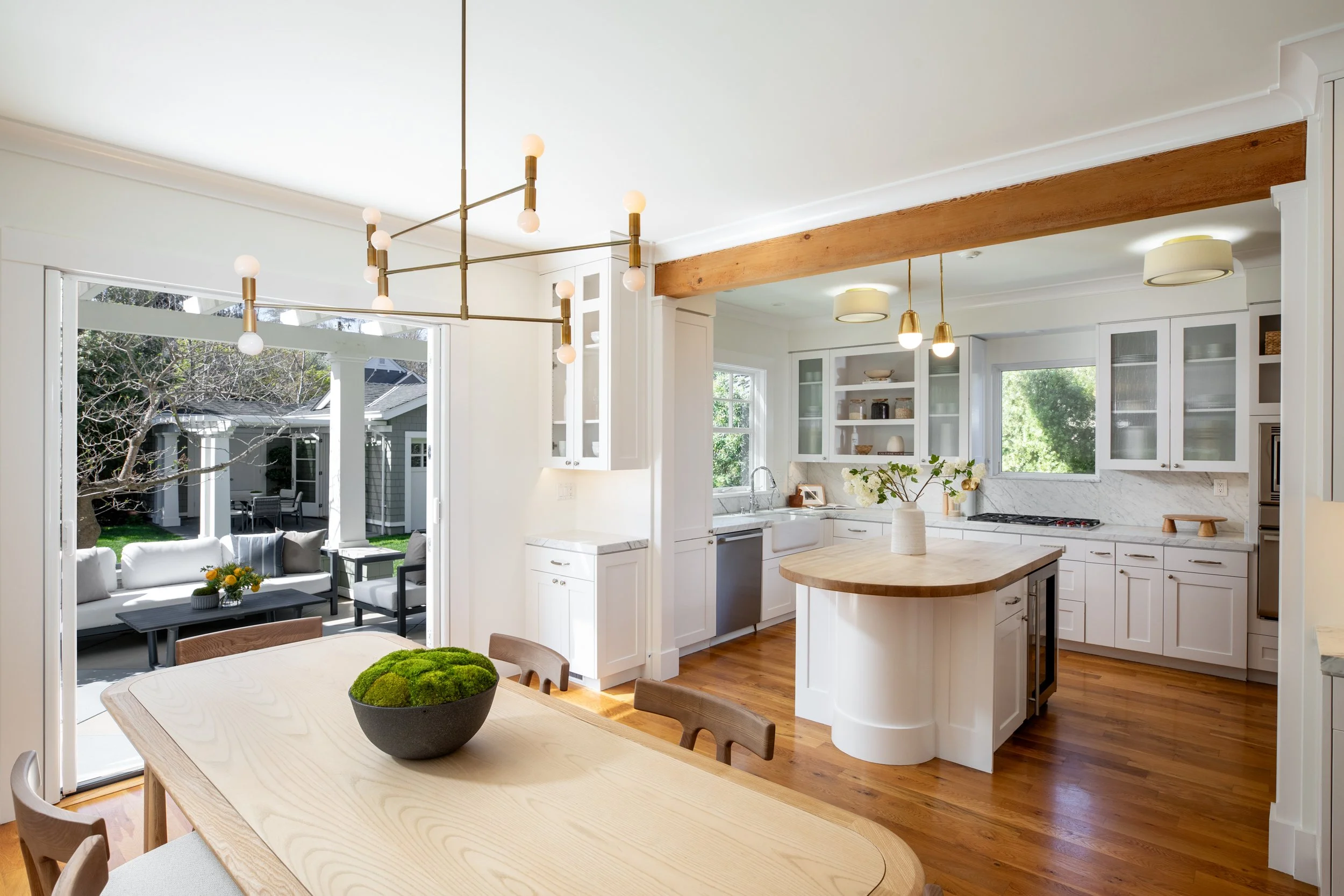 Bright kitchen with white cabinets, wooden floors, a central island, and a view into a sunlit outdoor patio with white furniture.