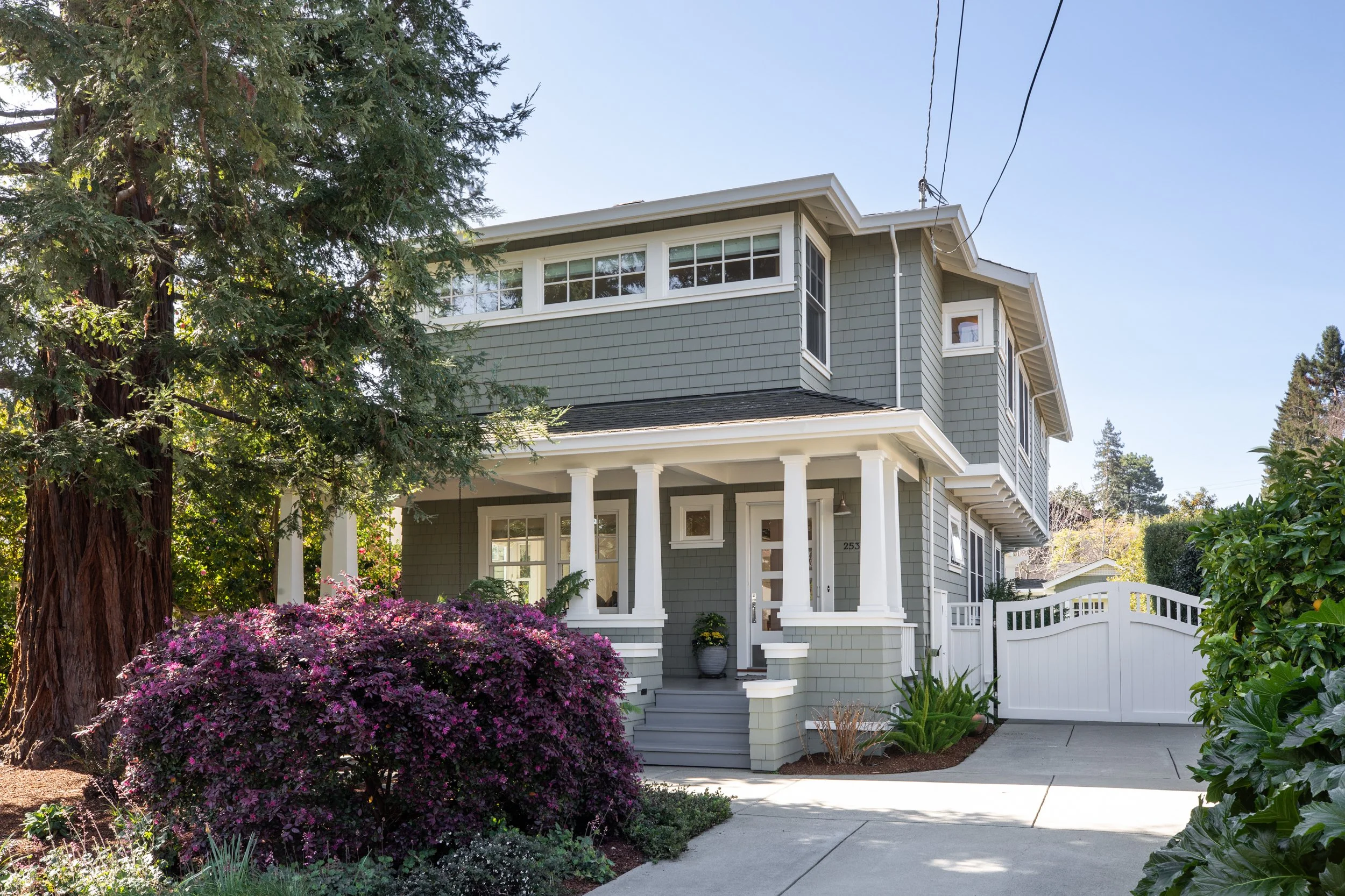 A grey, two-story house with white trim and a front porch with columns, surrounded by greenery and purple flowering bushes, with a white gate and sidewalk in front.