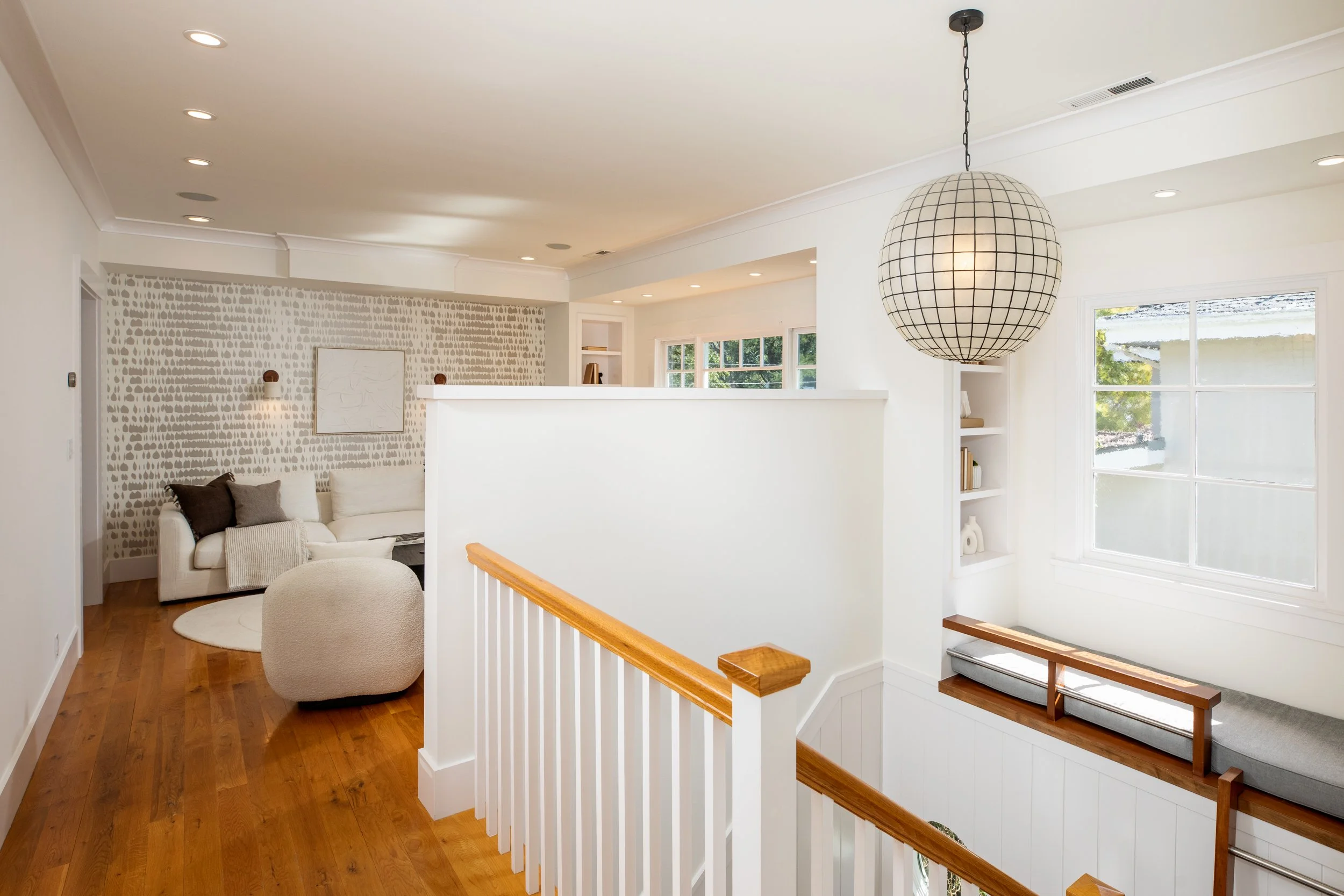 Bright, modern loft with white walls, hardwood floors, and large windows. There is a white sofa with dark pillows, a textured beige ottoman, built-in shelves, and a large spherical hanging light fixture.