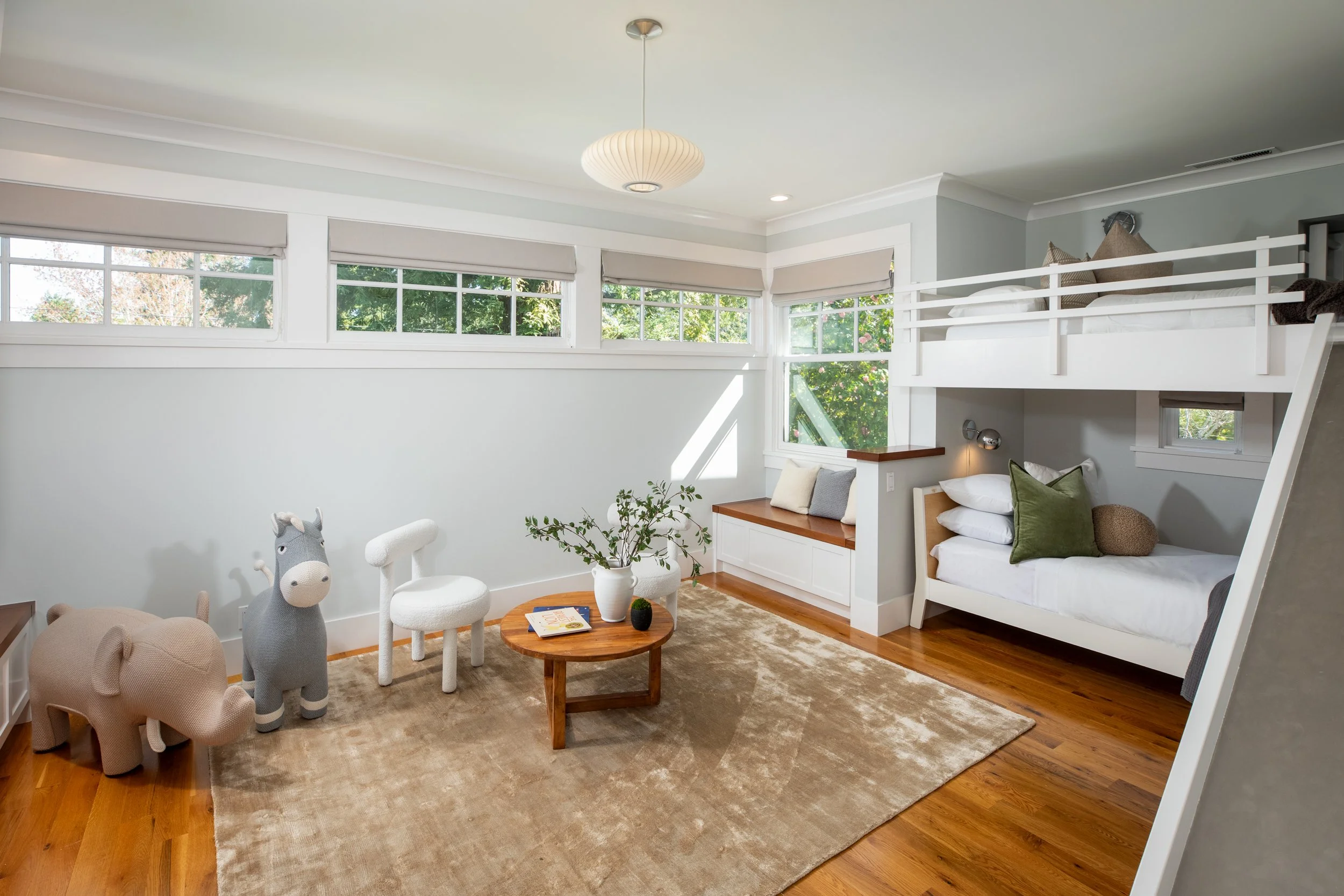 Children's bedroom featuring a loft bed with a slide, a sitting nook with pillows, a small round table with a plant, and plush animal toys on a beige rug.