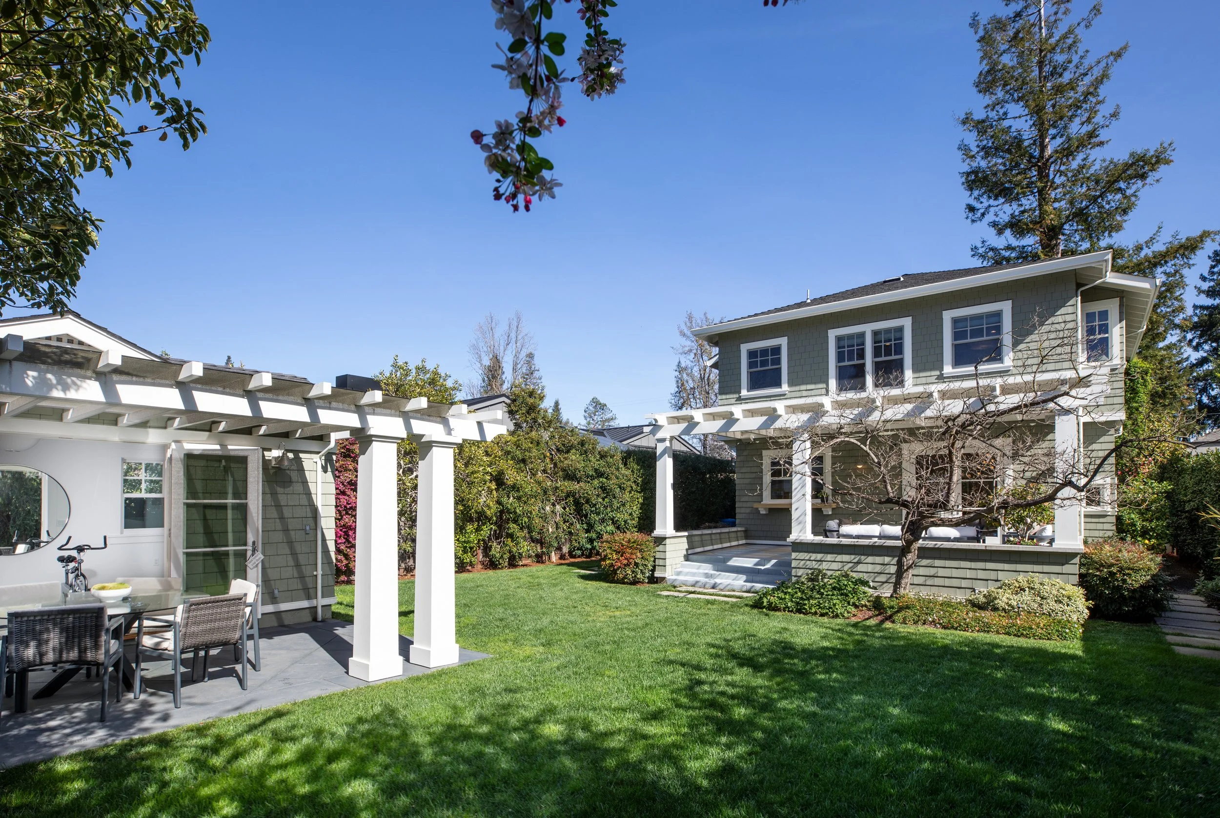 A backyard with a green lawn, a white pergola, and a two-story house with green siding, surrounded by trees and bushes, under a clear blue sky.