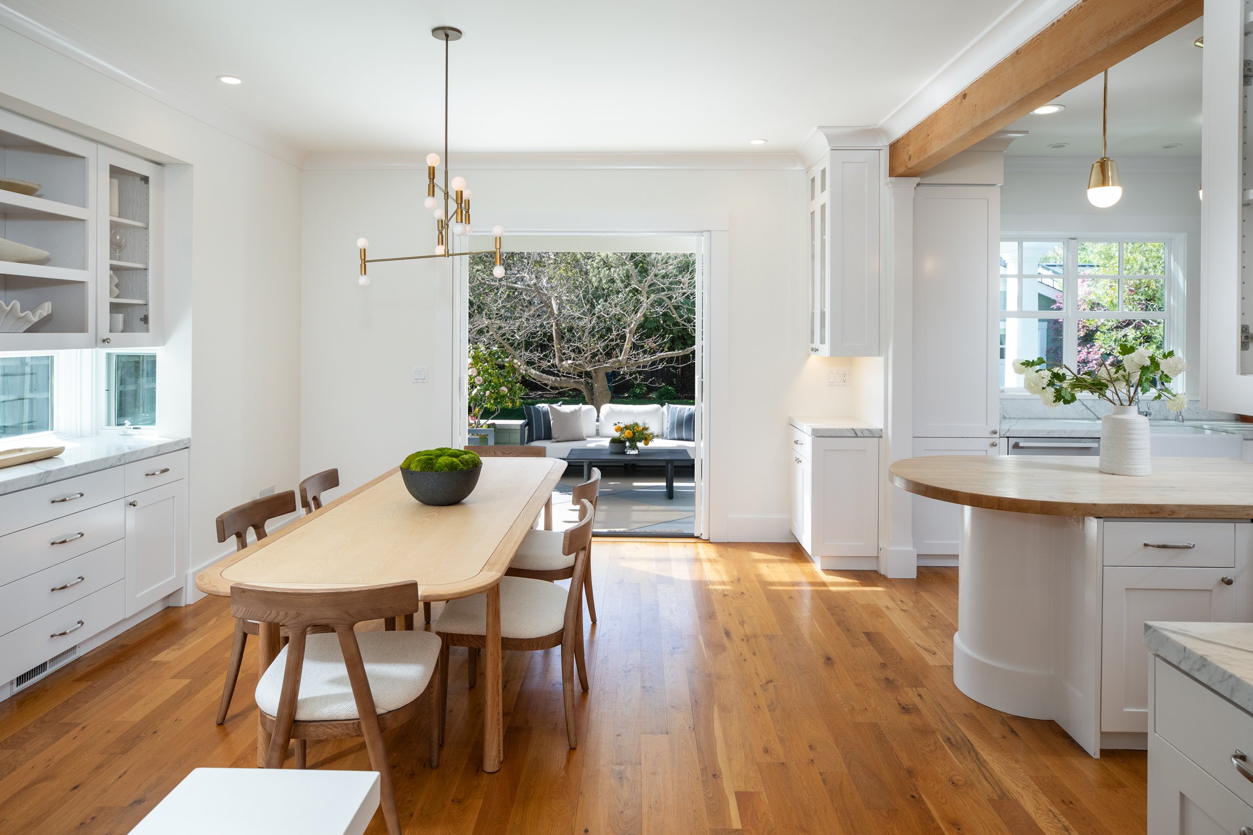 Bright, airy kitchen and dining area with white cabinetry, hardwood floors, a wooden dining table with six chairs, and sliding glass door leading to outdoor patio with seating