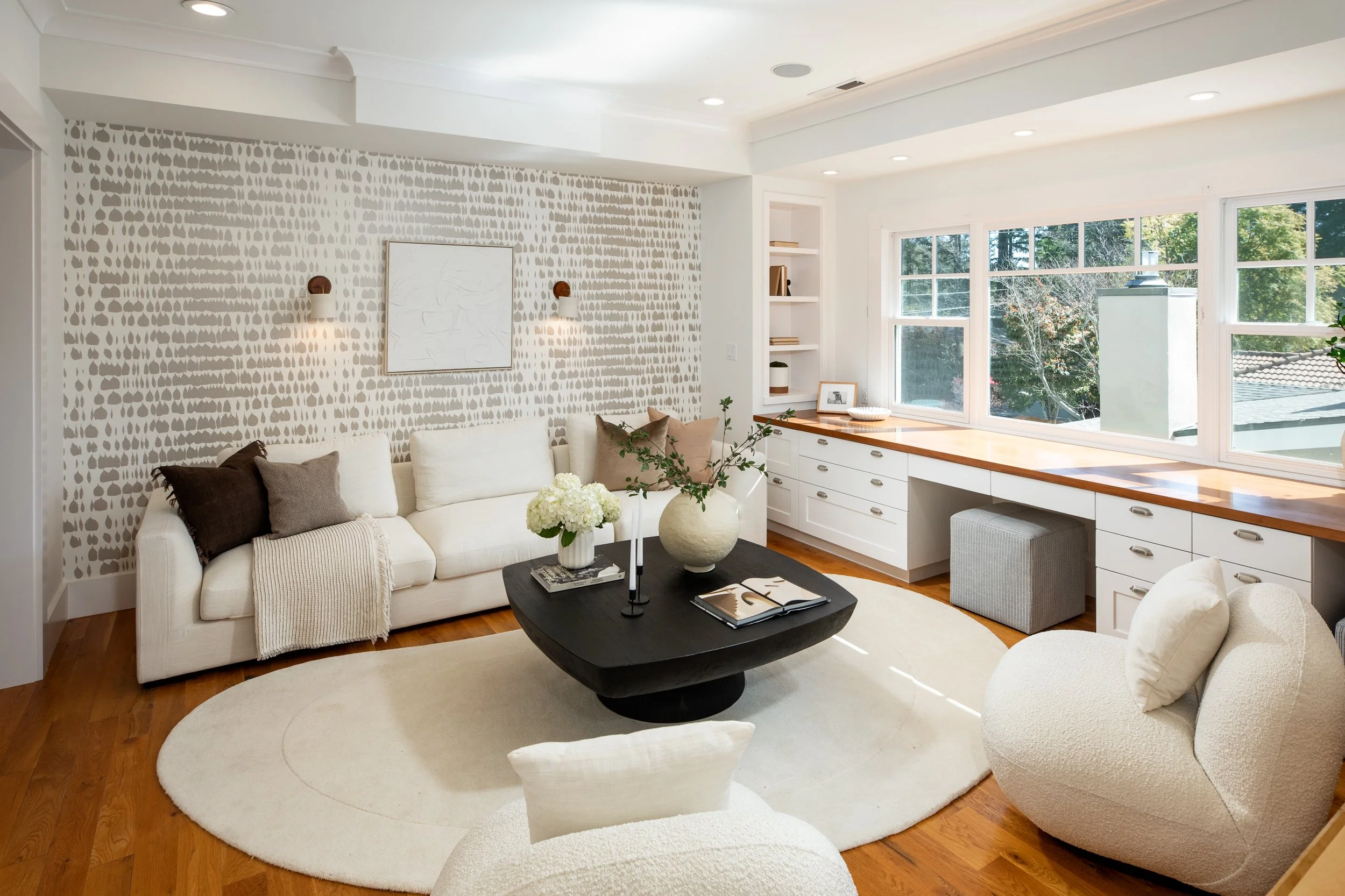 Bright loft family room room with white sofa, cream armchair, black coffee table, built-in white desk with drawers, large window with trees outside, decorative wall with abstract pattern, and minimal decor.