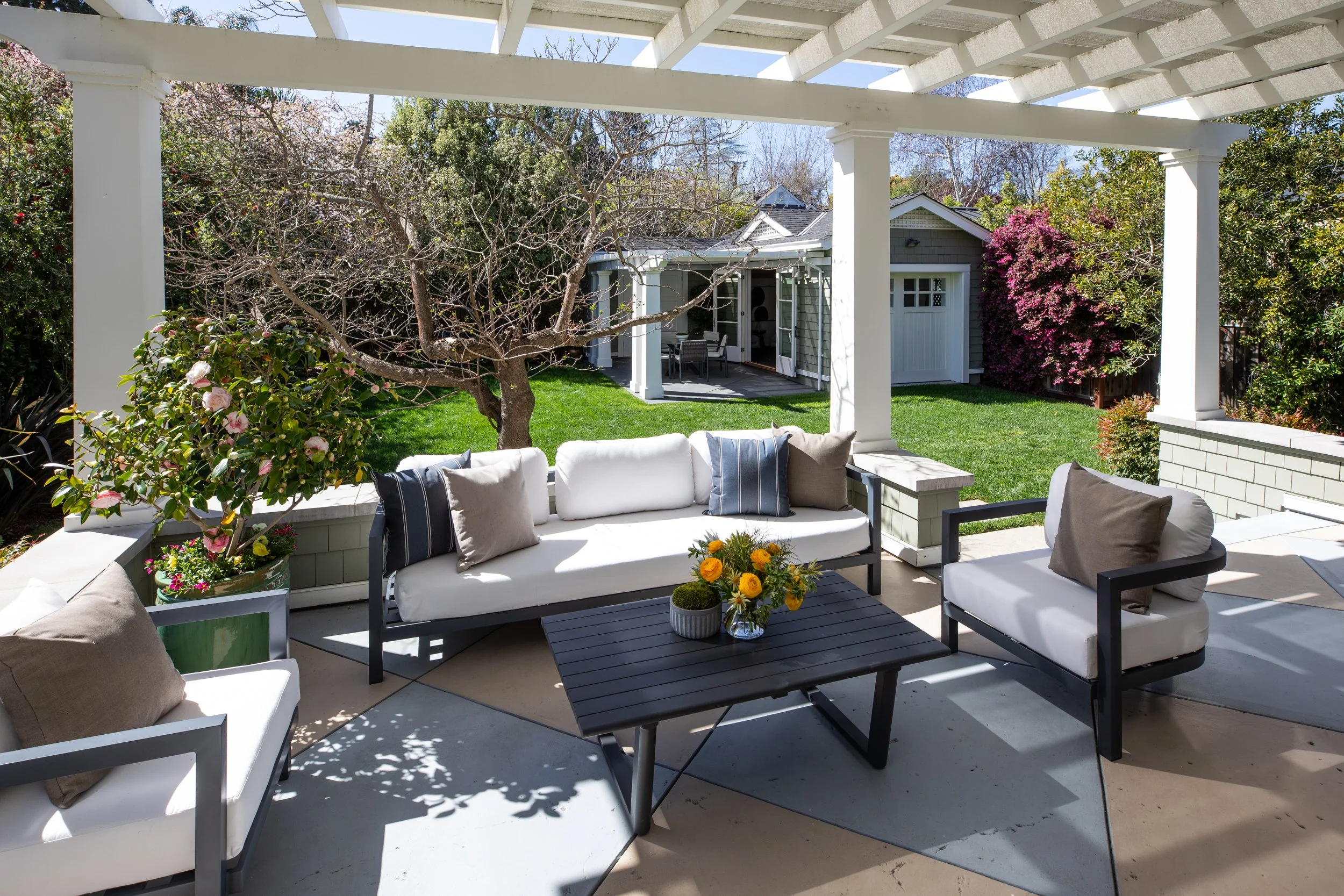 Patio with white cushioned outdoor furniture, a small black table with a flower arrangement, potted plant, green grass, trees, and a small shed in the background under a white pergola.