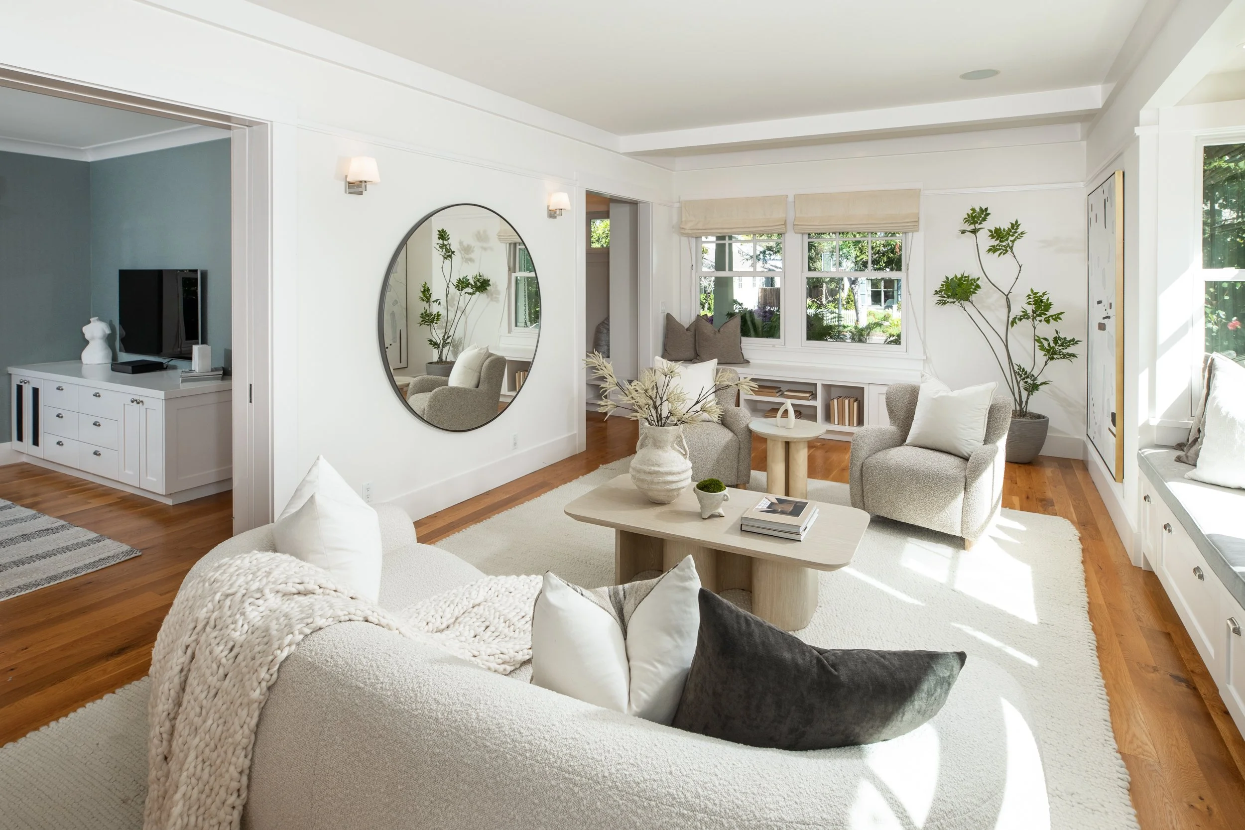 Bright living room with white walls, wooden floors, a large rounded sofa with white, black, and beige pillows, a beige throw blanket, and a wooden coffee table with books and decorative items. There are two armchairs, a built-in window seat with pill