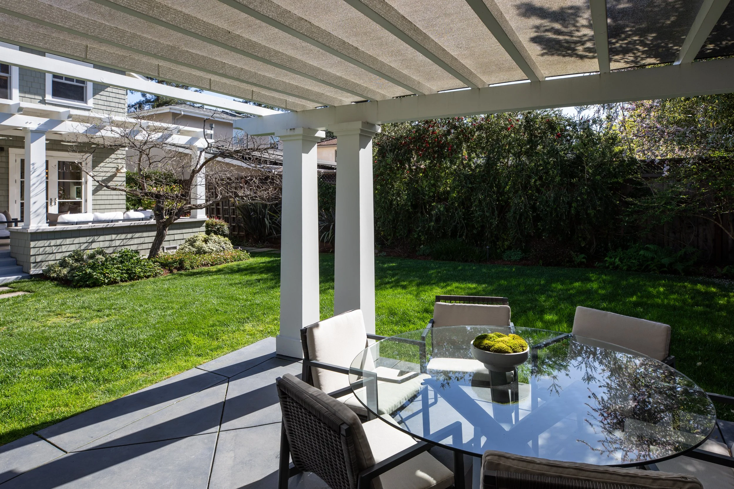 Backyard patio with a glass-top table and chairs, shaded under a pergola, with a lawn, garden, and a house in the background.