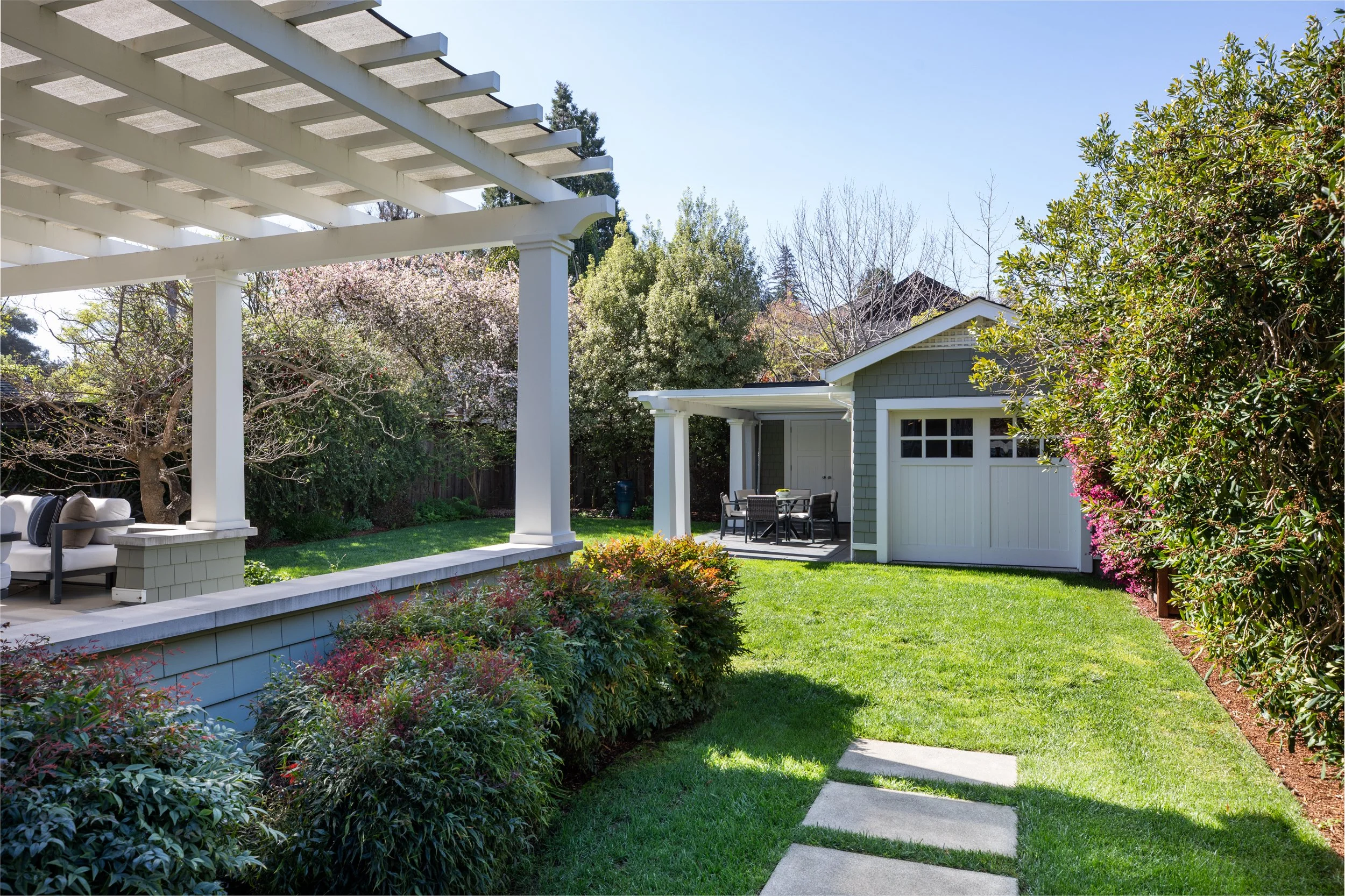A backyard with trimmed grass, a concrete stepping stone path, and a white patio with outdoor furniture under a pergola. There are trees and flowering bushes along the fence.