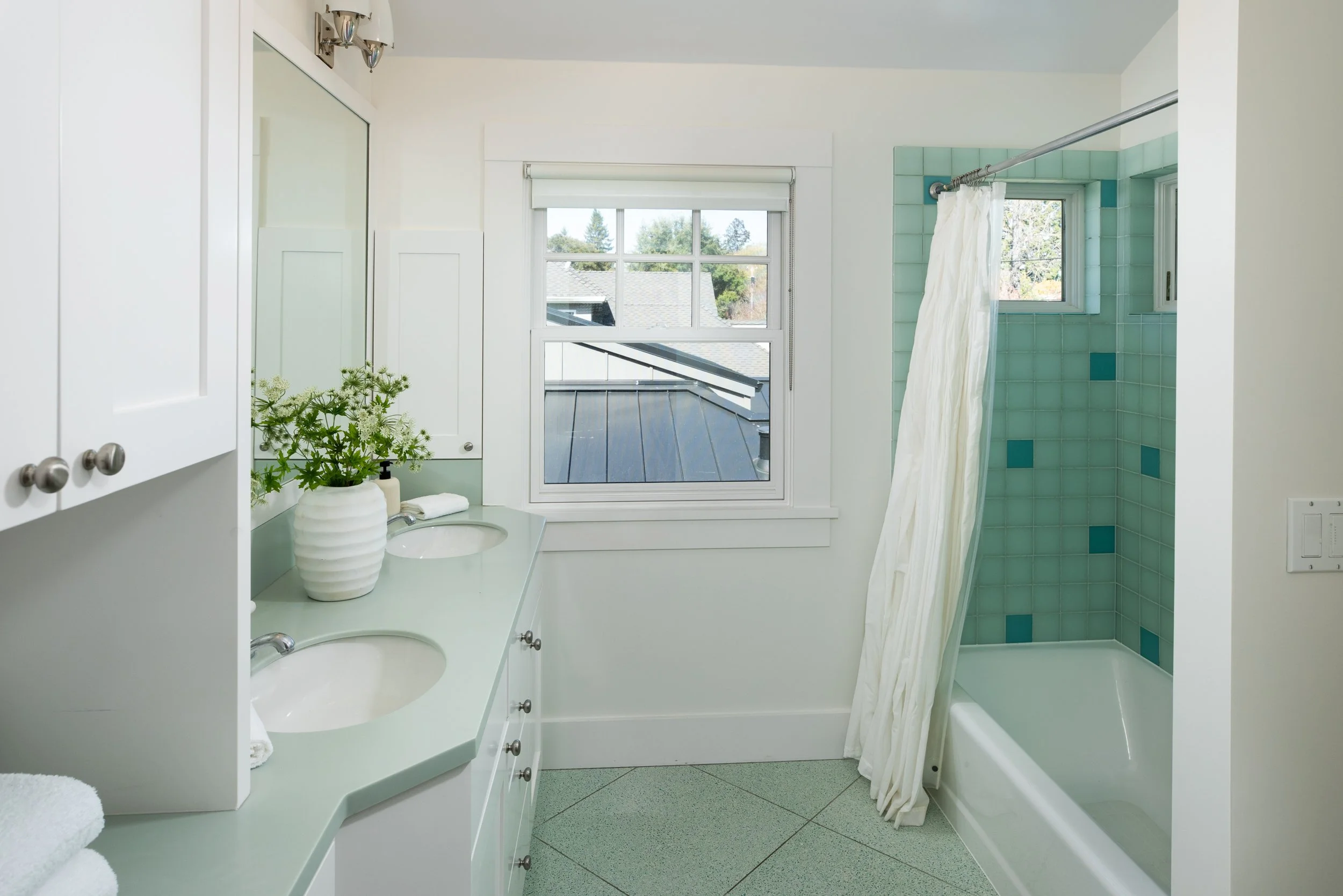 A bathroom featuring a dual sink vanity with a light green countertop, a white cabinet with silver knobs, a large mirror, a potted plant, a soap dispenser, and a towel. There is a window, a bathtub with a curtain, and aqua-colored tiles with darker a