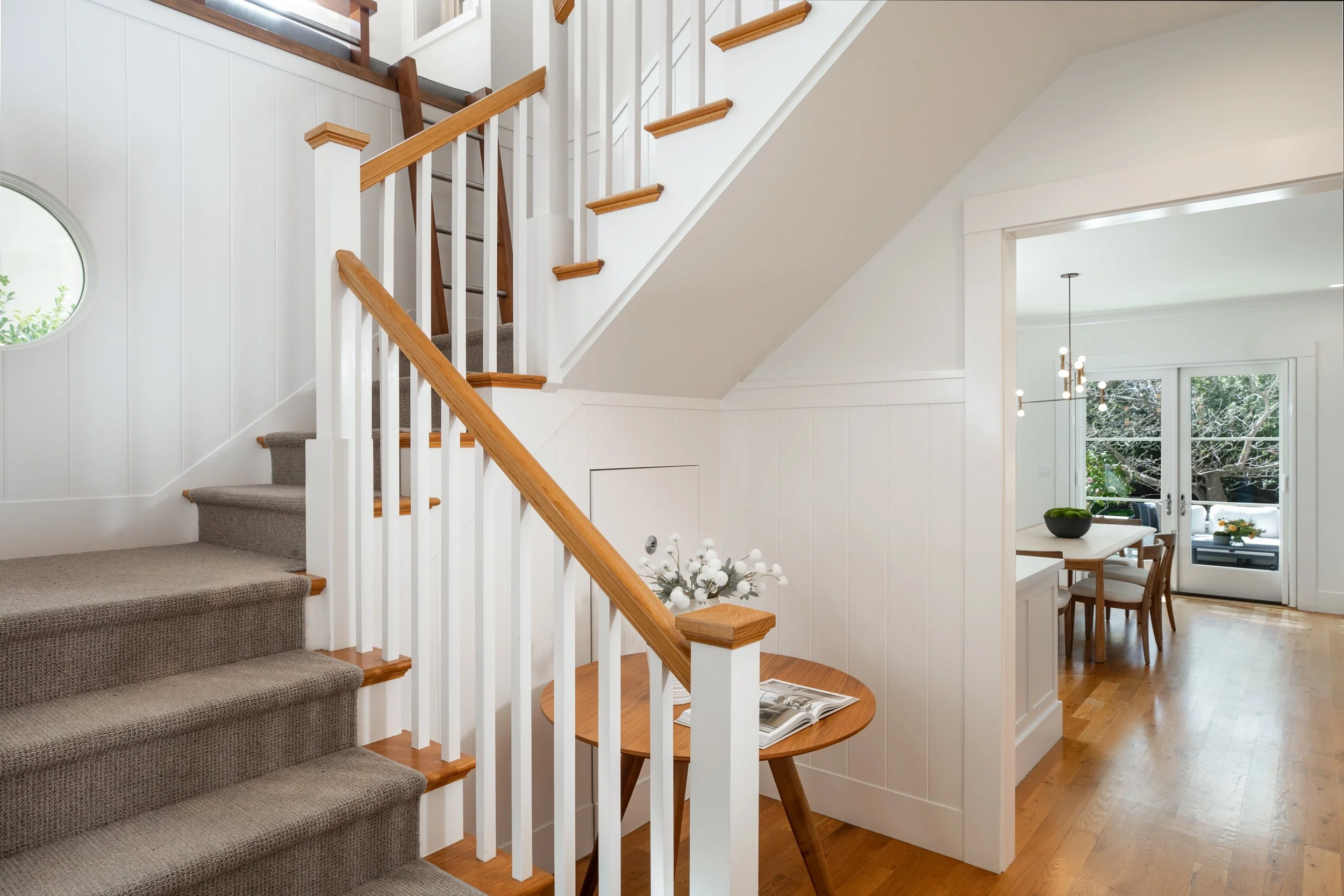 Interior of a home showing a staircase with carpeted steps, a small round wood table with a vase of white flowers and an open magazine, and a dining area with a table, chairs, and sliding glass doors leading outside.