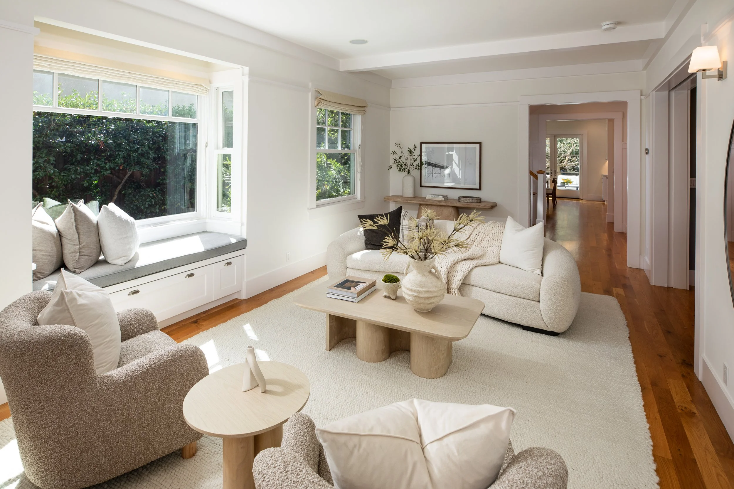 Bright living room with large window seat, white walls, light-colored sofa and armchairs, wooden floor, and minimal decor including vases, books, and artwork.