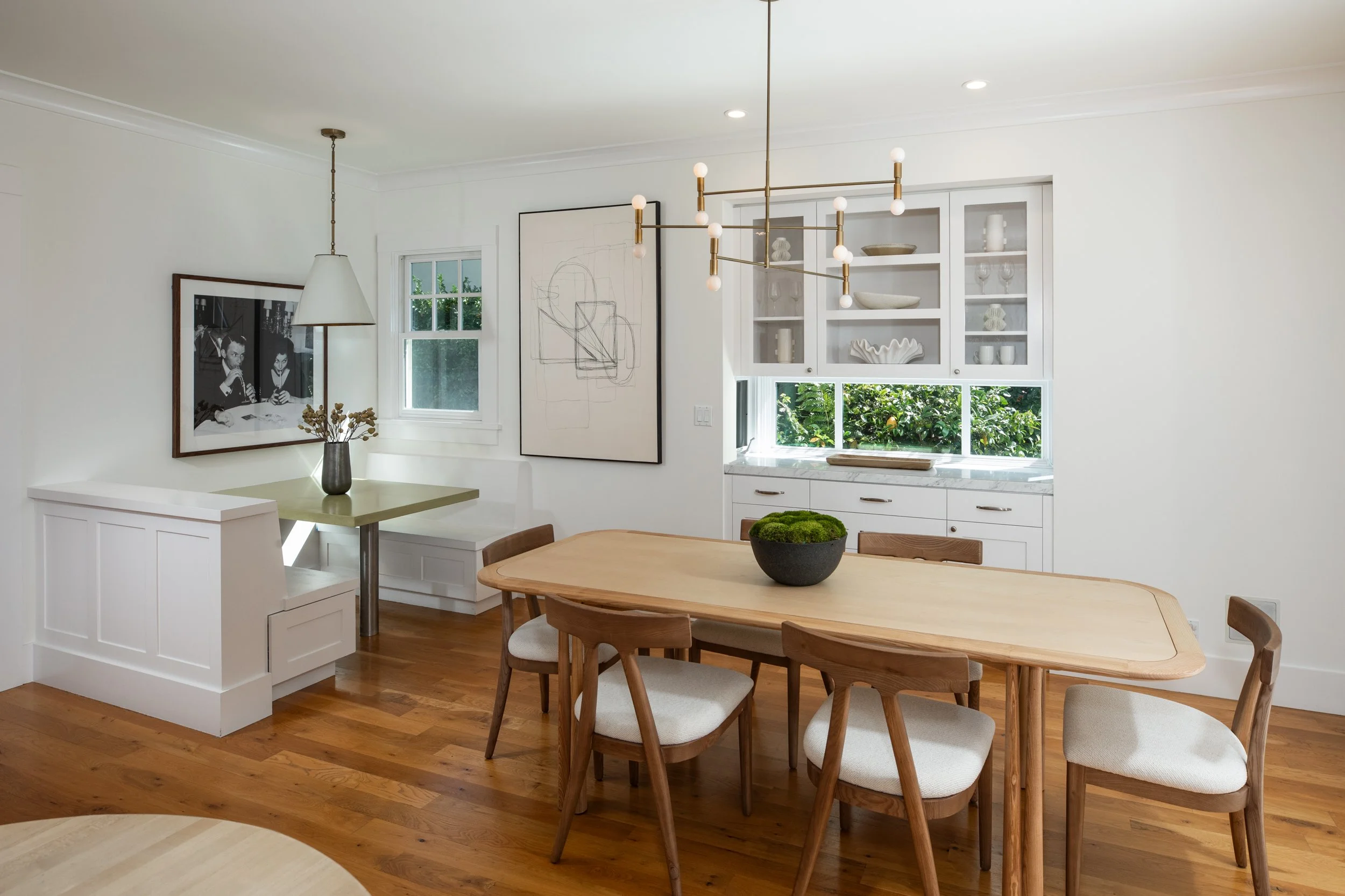 A modern dining room with a wooden table and six matching chairs, a black bowl with green moss as a centerpiece, white cabinetry with glass doors, a decorative chandelier, and artwork on the white walls.