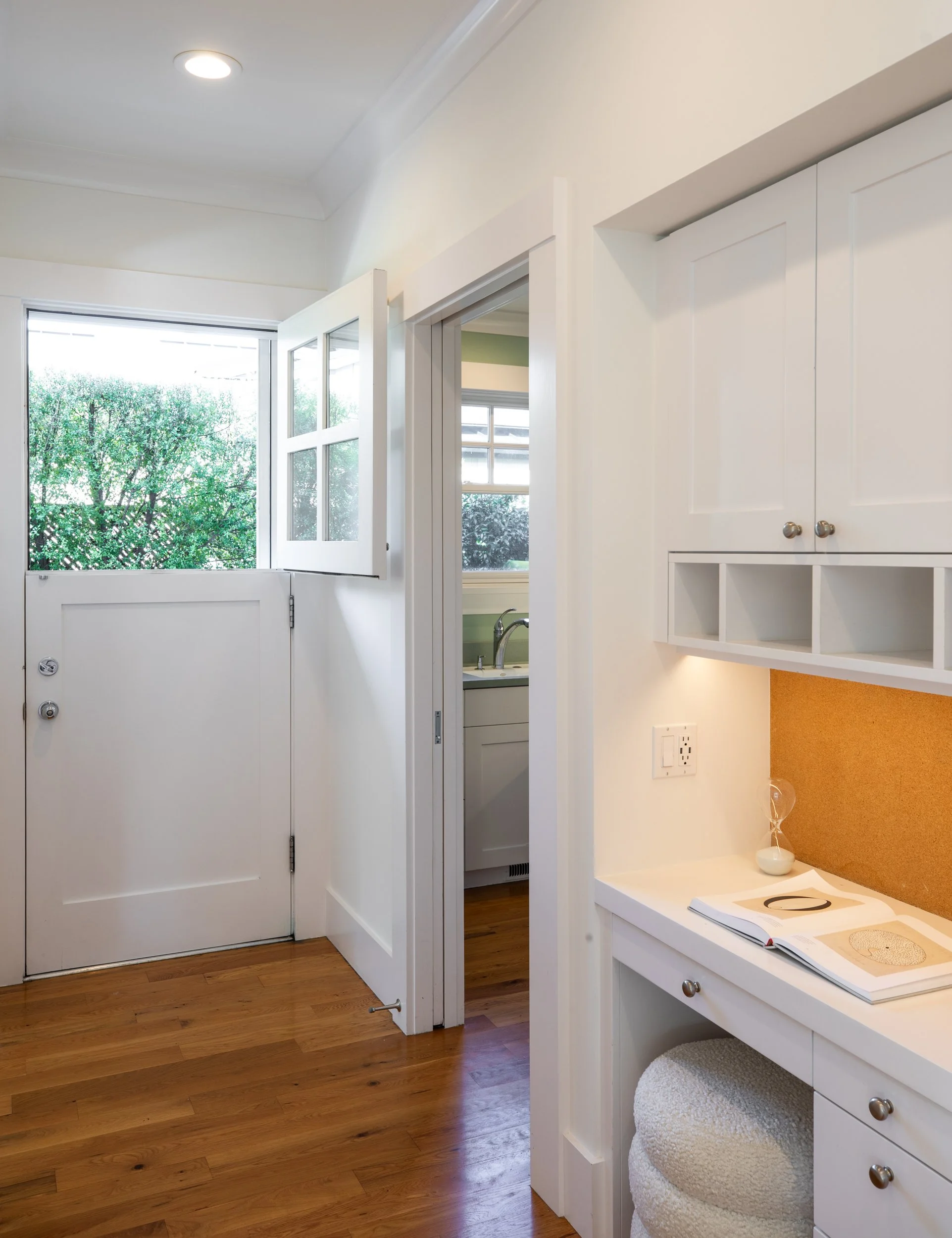 A bright entryway with an open white door leading outside, a small kitchenette visible through an open doorway, hardwood floors, and a white built-in desk with open shelves and a corkboard.