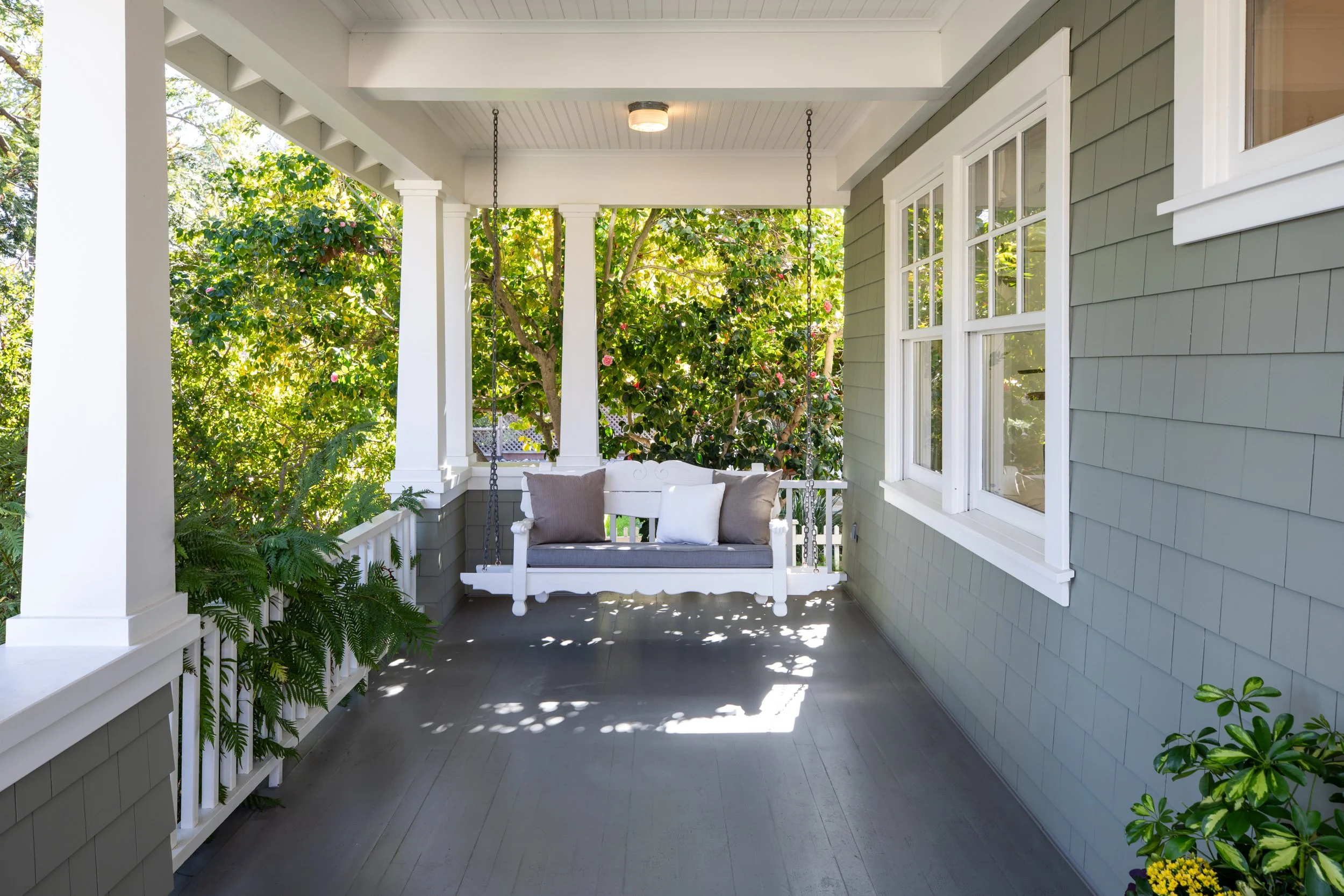 Porch with a white swing with three pillows, surrounded by greenery and windows of a house.