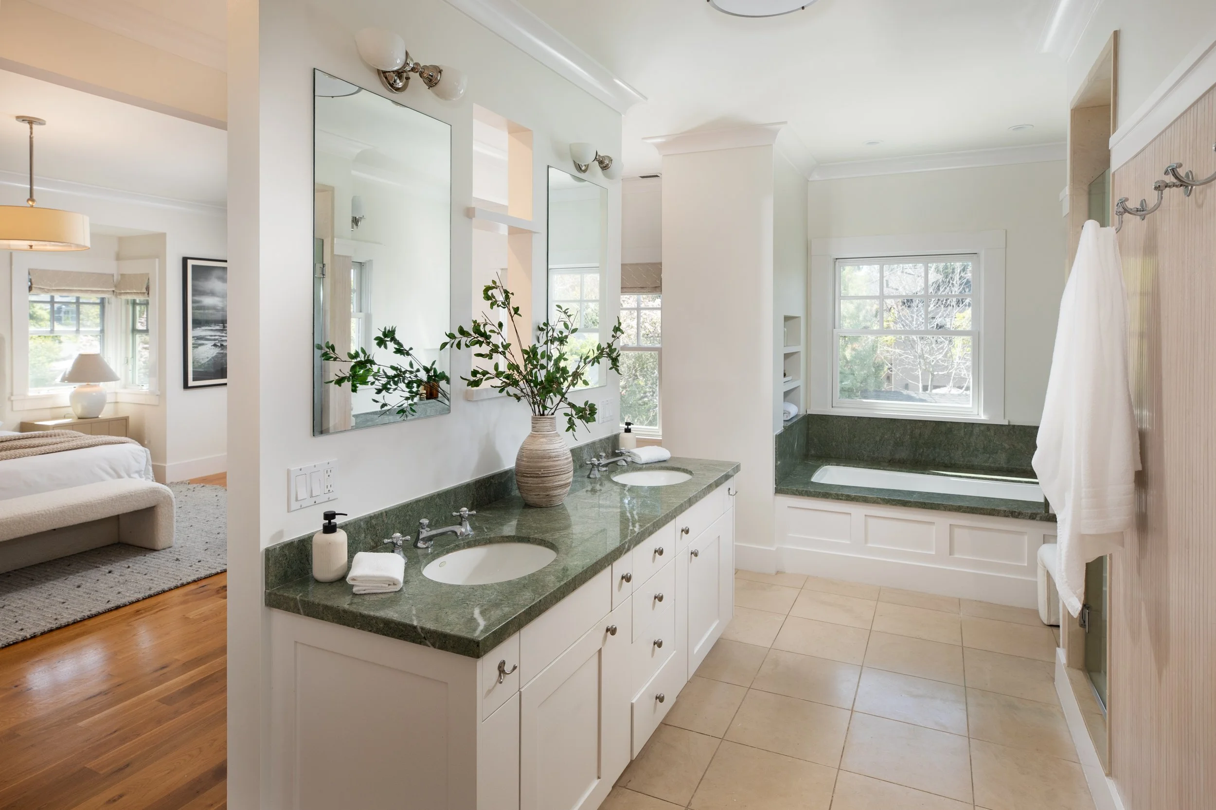 Bathroom with double sink vanity with green marble countertop, a vase with green branches, two mirrors, and a bathtub with a window above it.