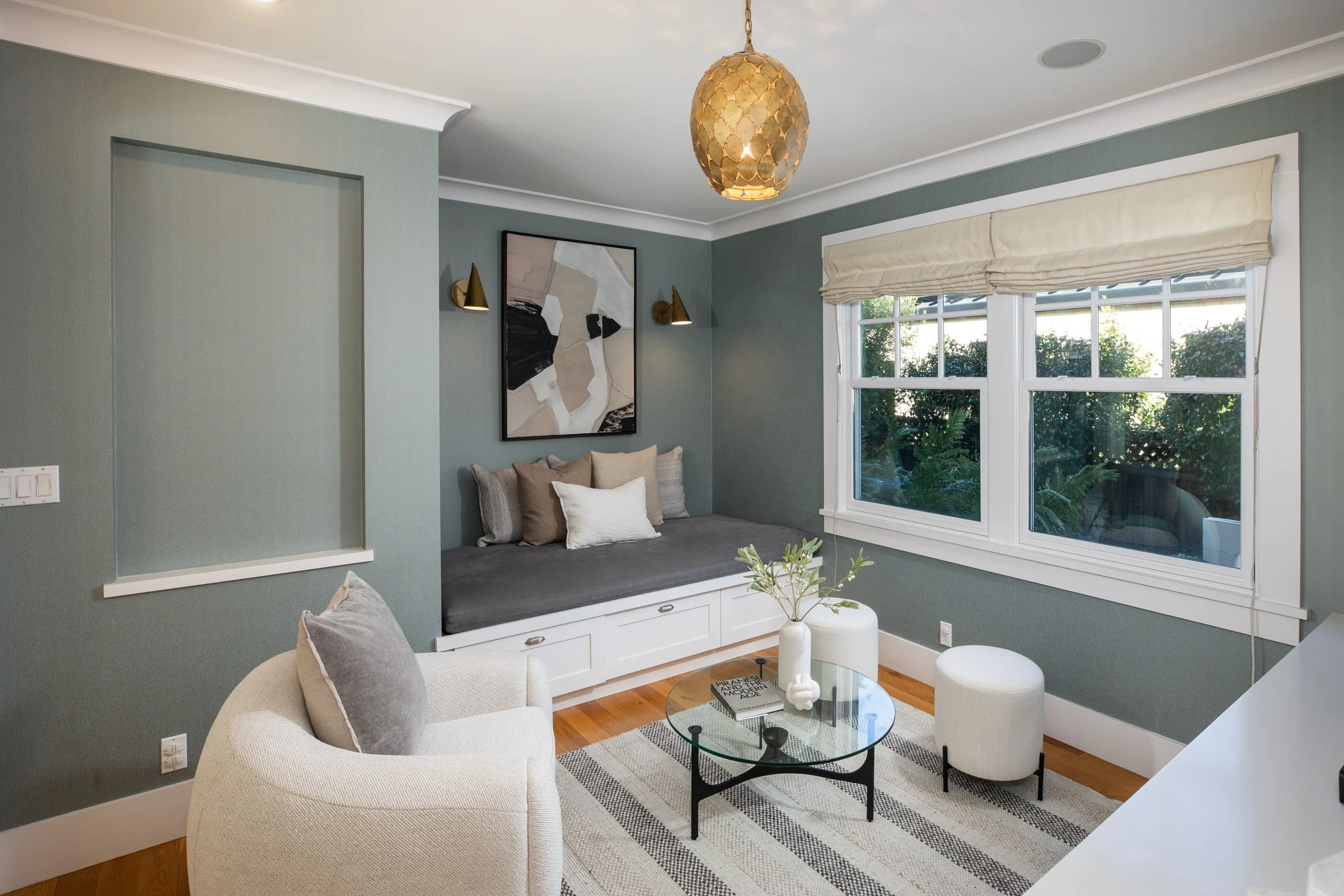 A cozy modern den with a built-in window seat, gray and beige accents, a glass coffee table, and a large window with a view of greenery outside.