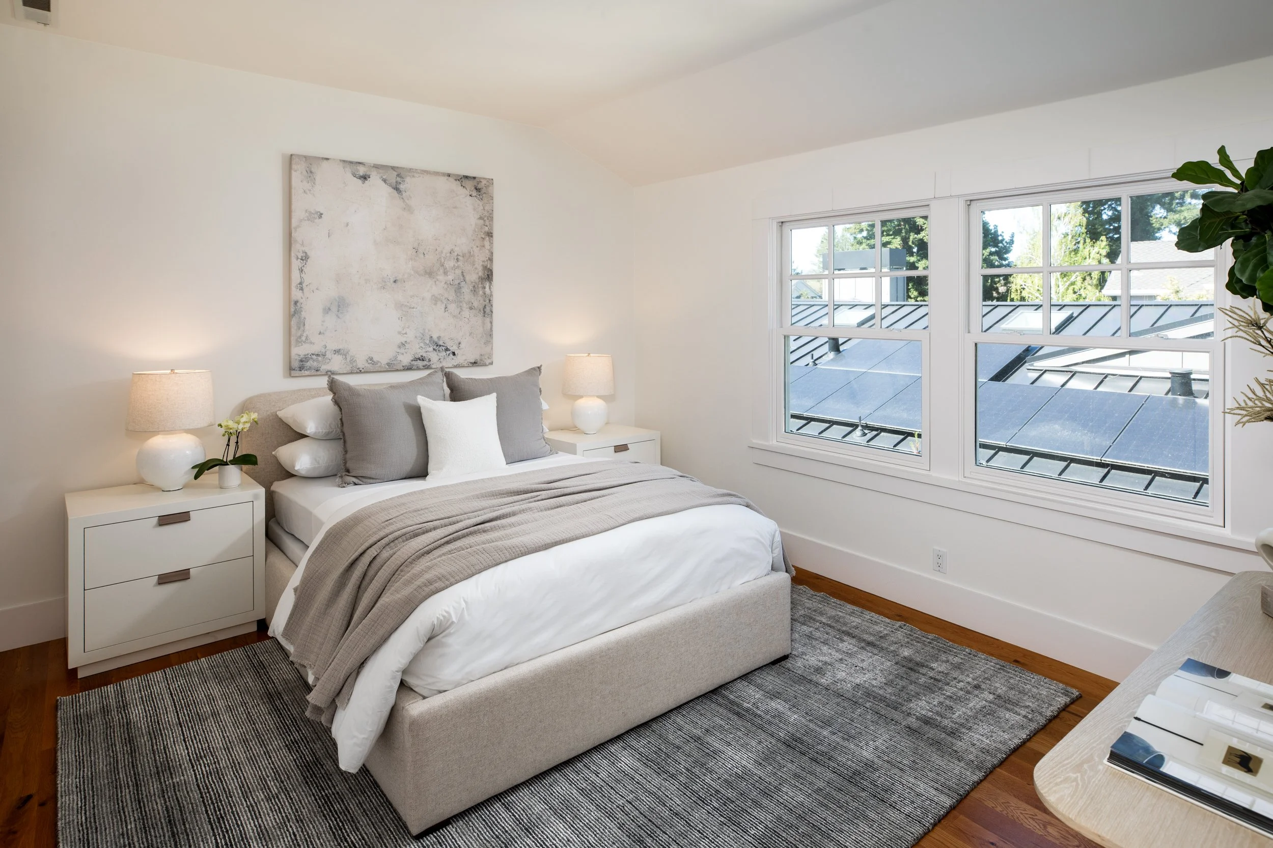 A modern bedroom with a bed, gray and white bedding, two nightstands with lamps, and a large window showing rooftops and trees outside.