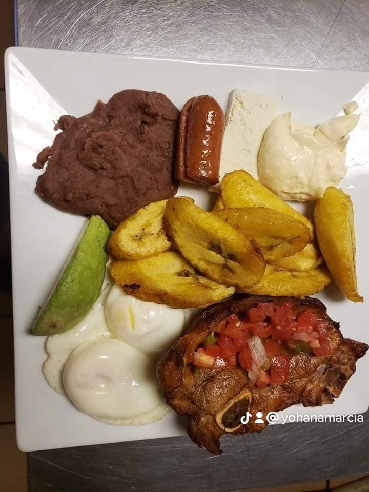 Plate of Mexican food with refried beans, grilled sausage, queso fresco, guacamole, fried plantains, sour cream, grilled pork chop with salsa, and a slice of cucumber.