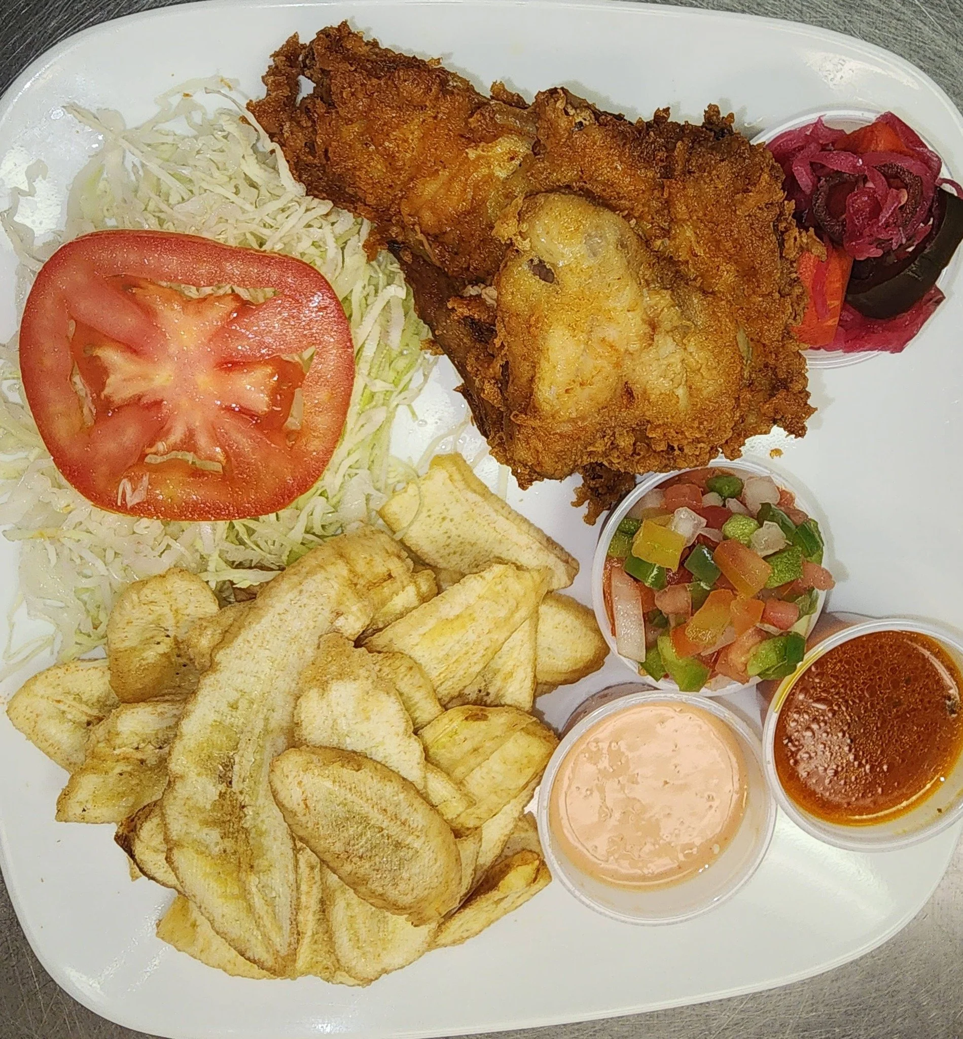 Plate with fried fish fillet, potato chips, sliced tomato, shredded cabbage, pickled vegetables, and three small cups of dipping sauces.