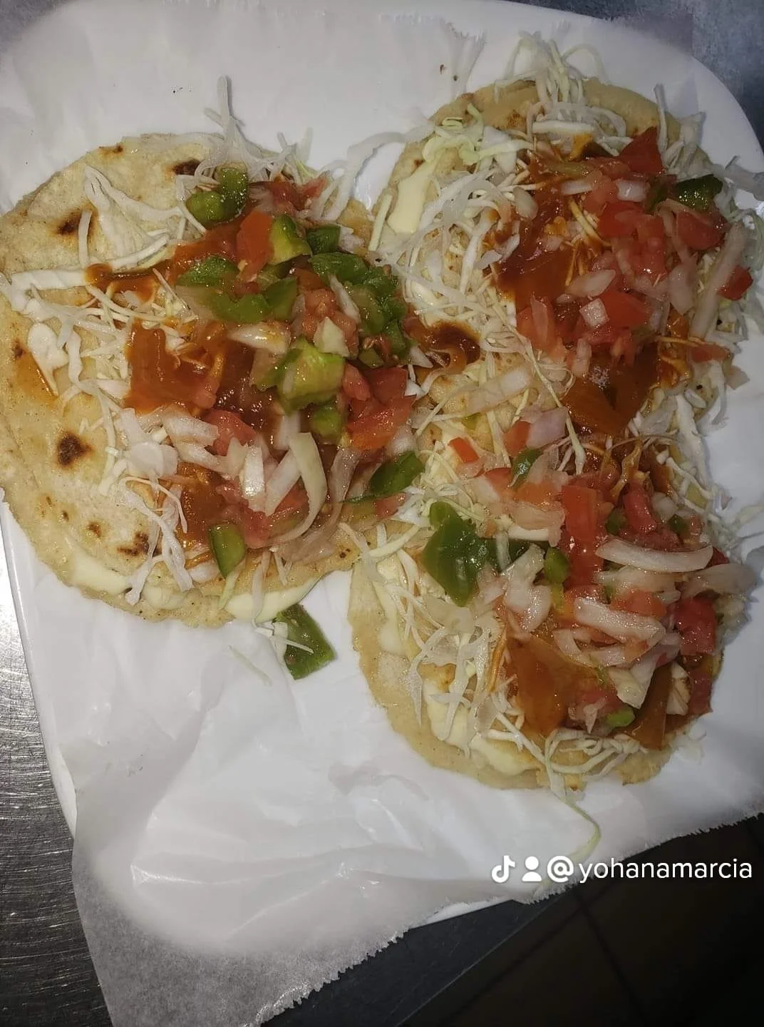Three tostadas topped with shredded cheese, chopped tomatoes, diced green peppers, shredded lettuce, and hot sauce, on a white paper-lined tray.