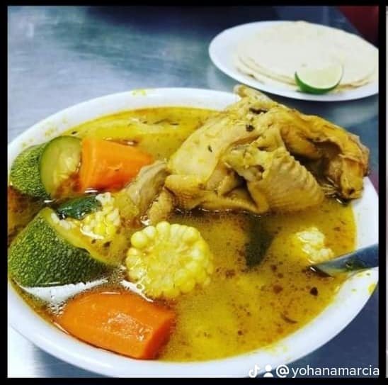 Soup with chicken, zucchini, carrots, corn, and other vegetables in a white bowl, with a side of tortillas and lime wedge in the background.