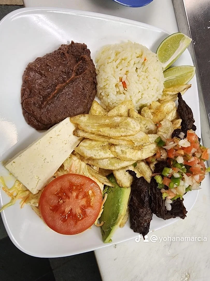 Plate of Mexican meal with grilled beef, rice, fried plantains, sliced tomato, cheese, guacamole, refried black beans, and lime wedges.