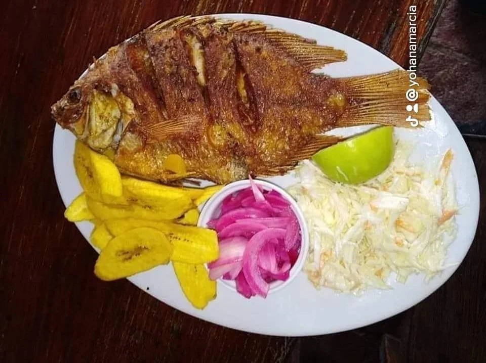 Grilled whole fish with plantain chips, pickled onions, shredded cabbage, a lime wedge, and a lime half on a white plate.