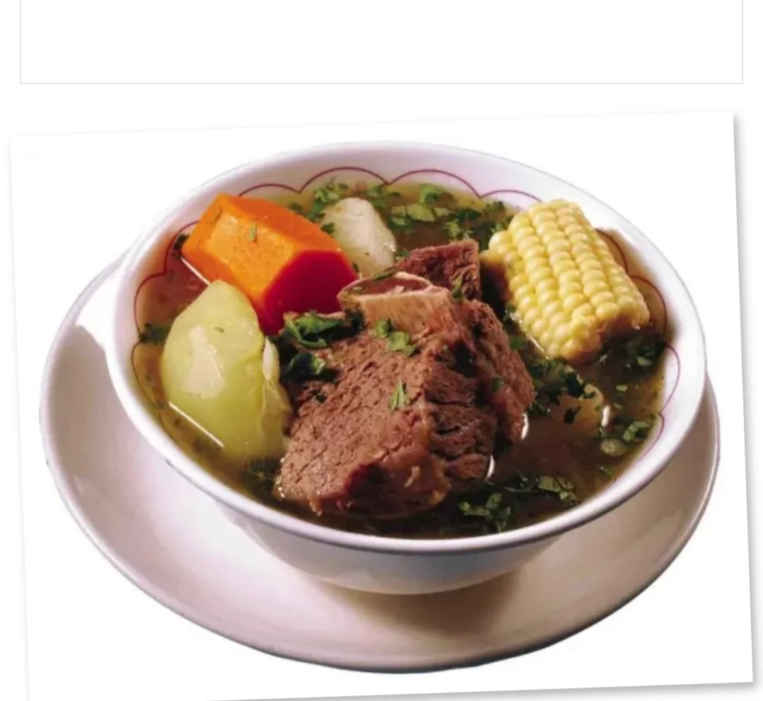 Bowl of beef soup with potatoes, corn on the cob, squash, and herbs on a white plate.