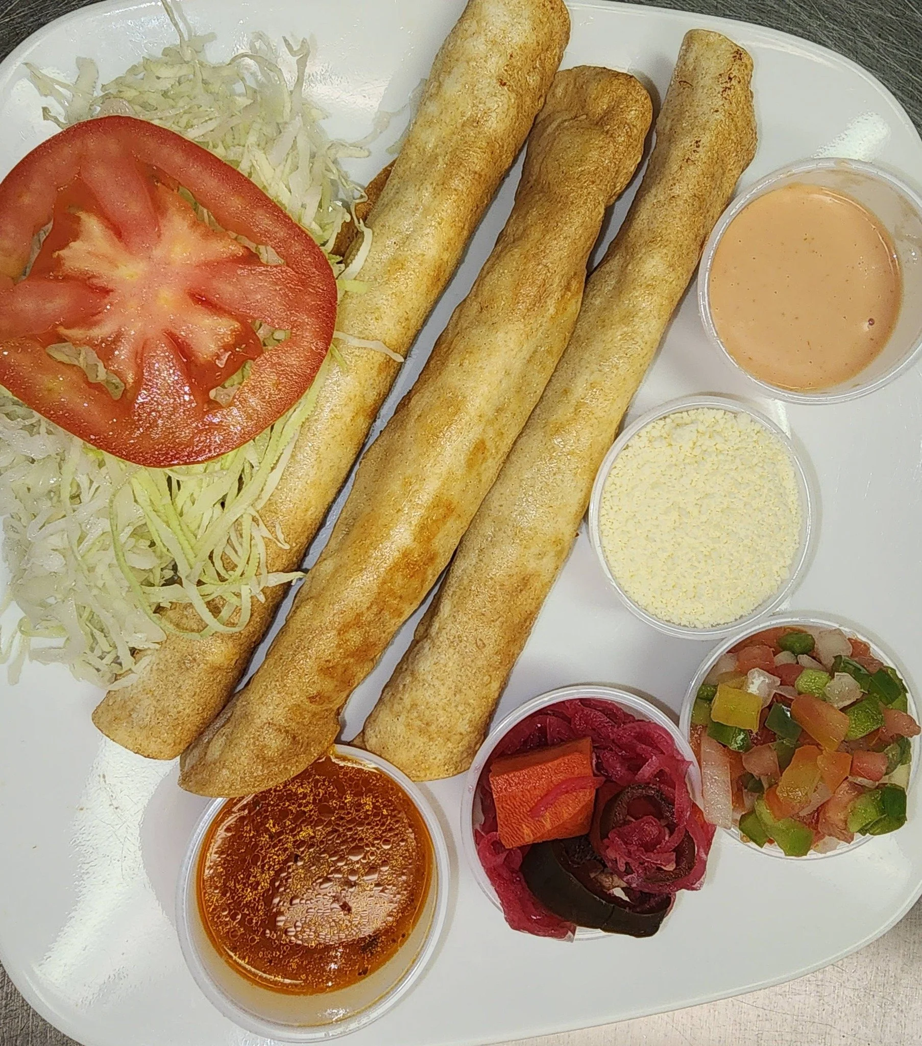 Plate of Mexican taquitos with shredded lettuce and tomato, served with various sauces, cheese, and pickled vegetables.