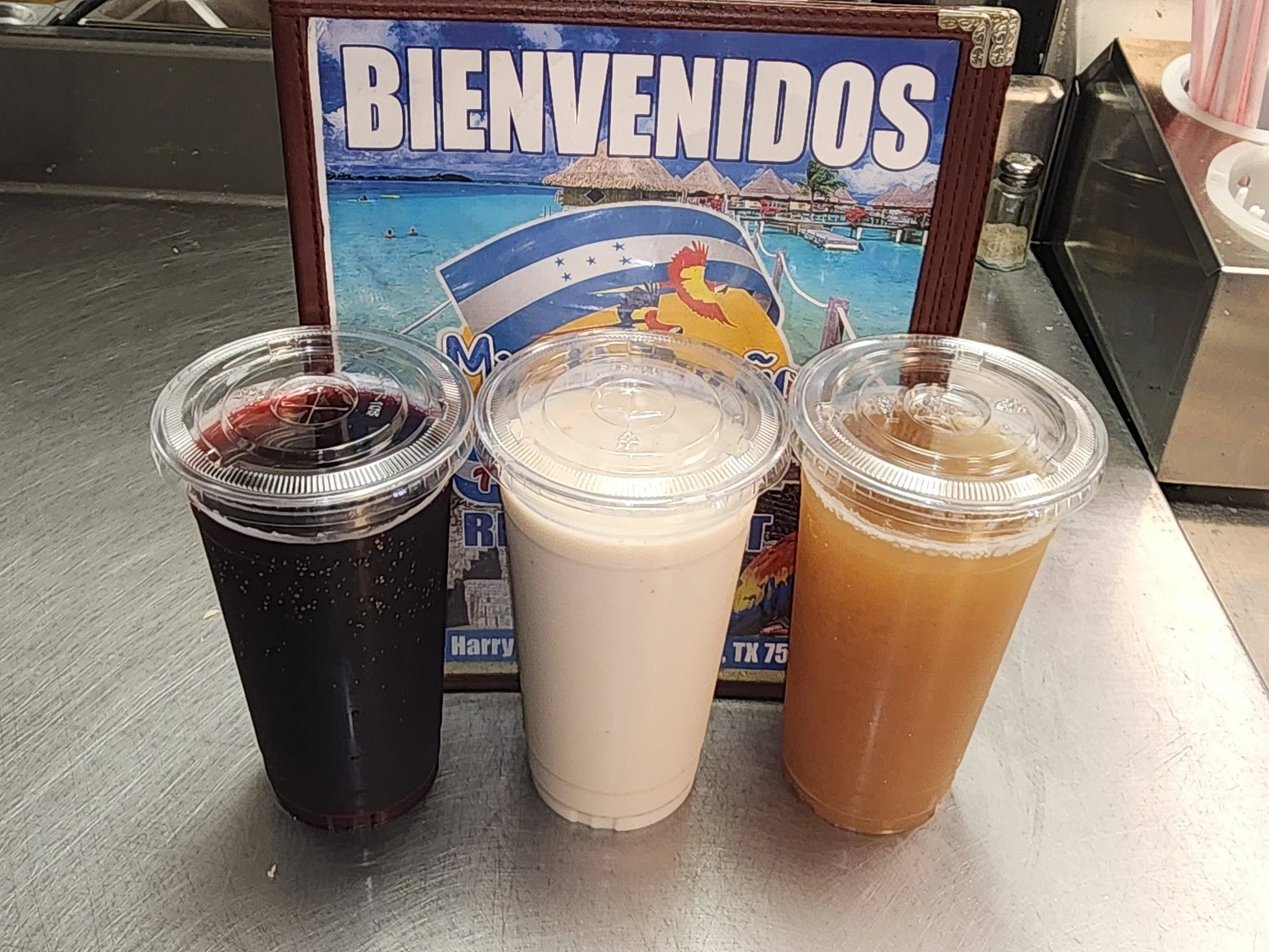Three large plastic cups of beverages on a table with a welcome sign behind them. The drinks include a dark soda, a milkshake, and an orange juice or tea.