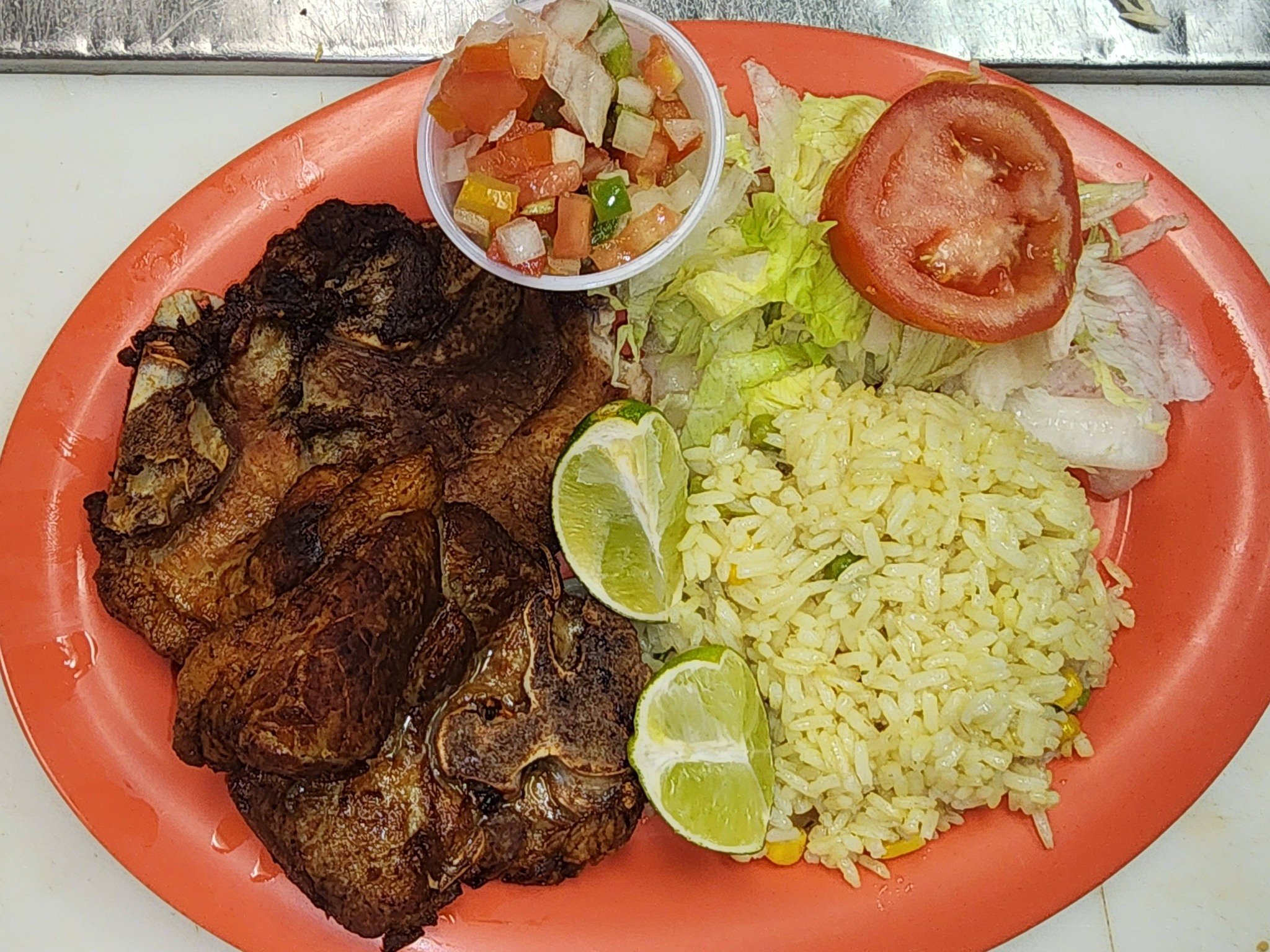 A plate of fried chicken, yellow rice, sliced tomato, lettuce, lime wedges, and a small cup of chopped vegetables or salsa.
