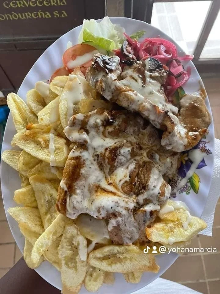 Plate of pork with white sauce, served with potato chips and salad with tomato, lettuce, and pickled pink onions.