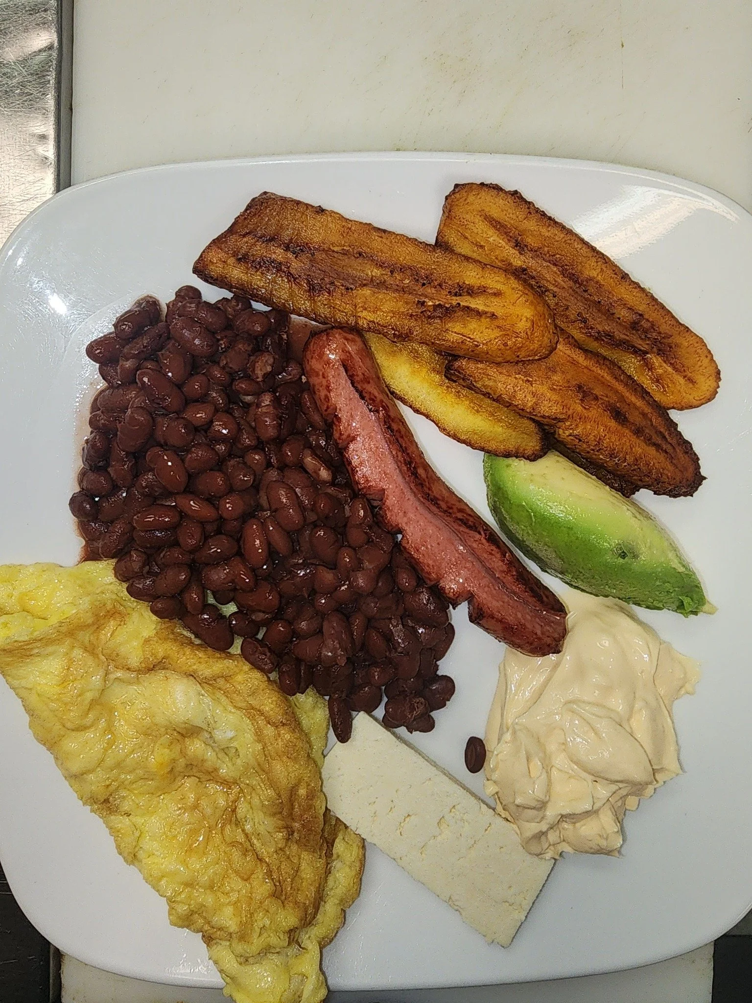 Fried plantains, baked beans, fried egg, sausage, sliced avocado, slices of white cheese, and a dollop of mayonnaise on a white plate.