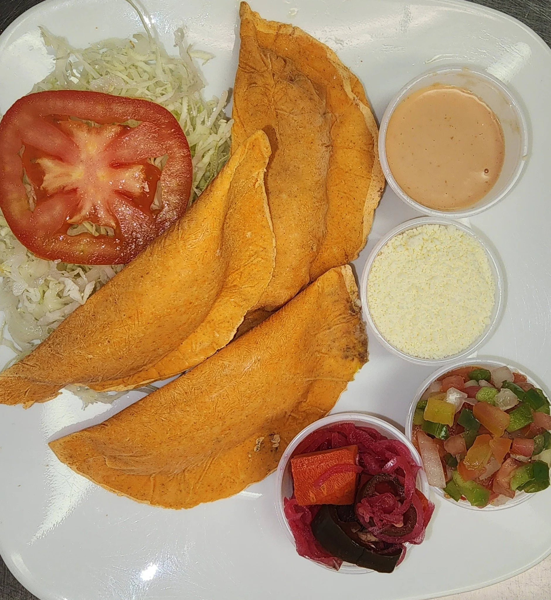Fried fish fillets with shredded lettuce and a slice of tomato, served with dipping sauces, grated cheese, and side salads of pickled vegetables and pico de gallo on a white plate.